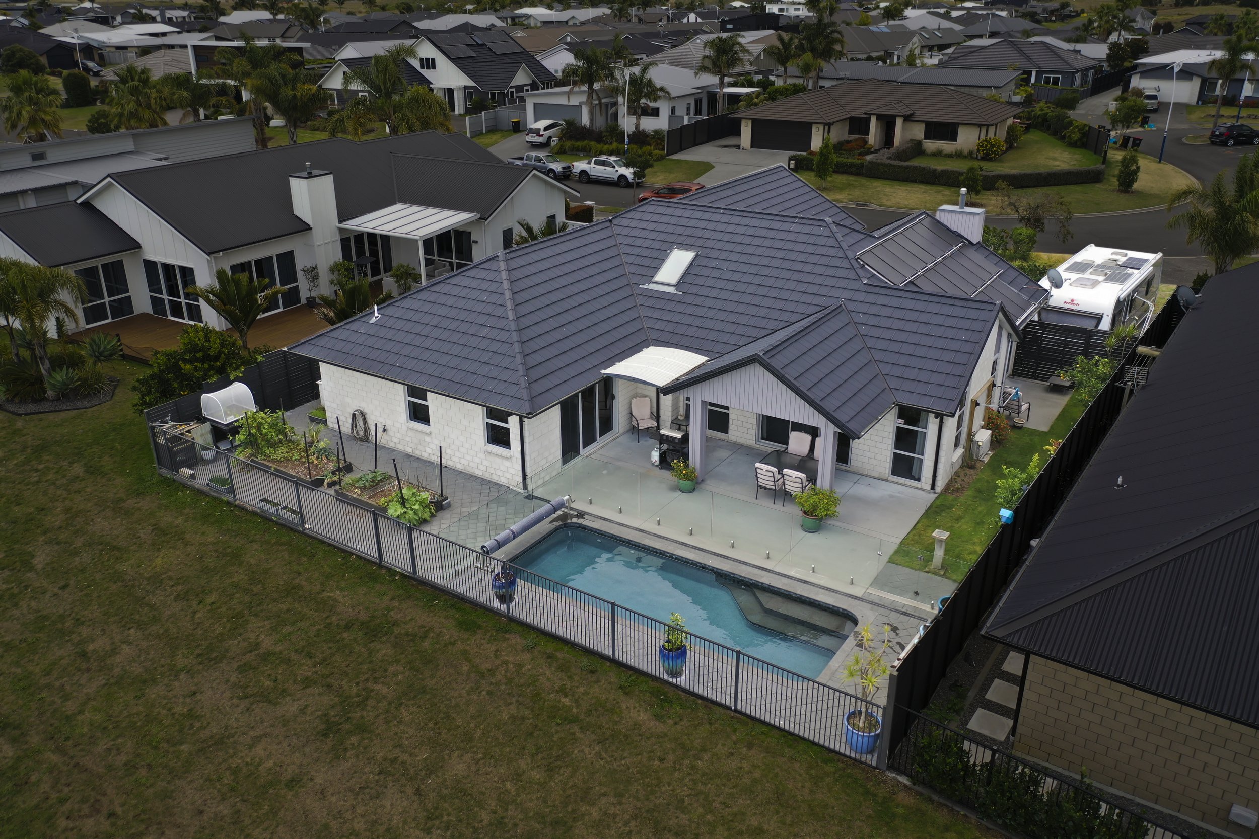 Aerial view of a modern suburban house with a backyard pool, patio, garden, and neighboring houses in a neighborhood with palm trees.