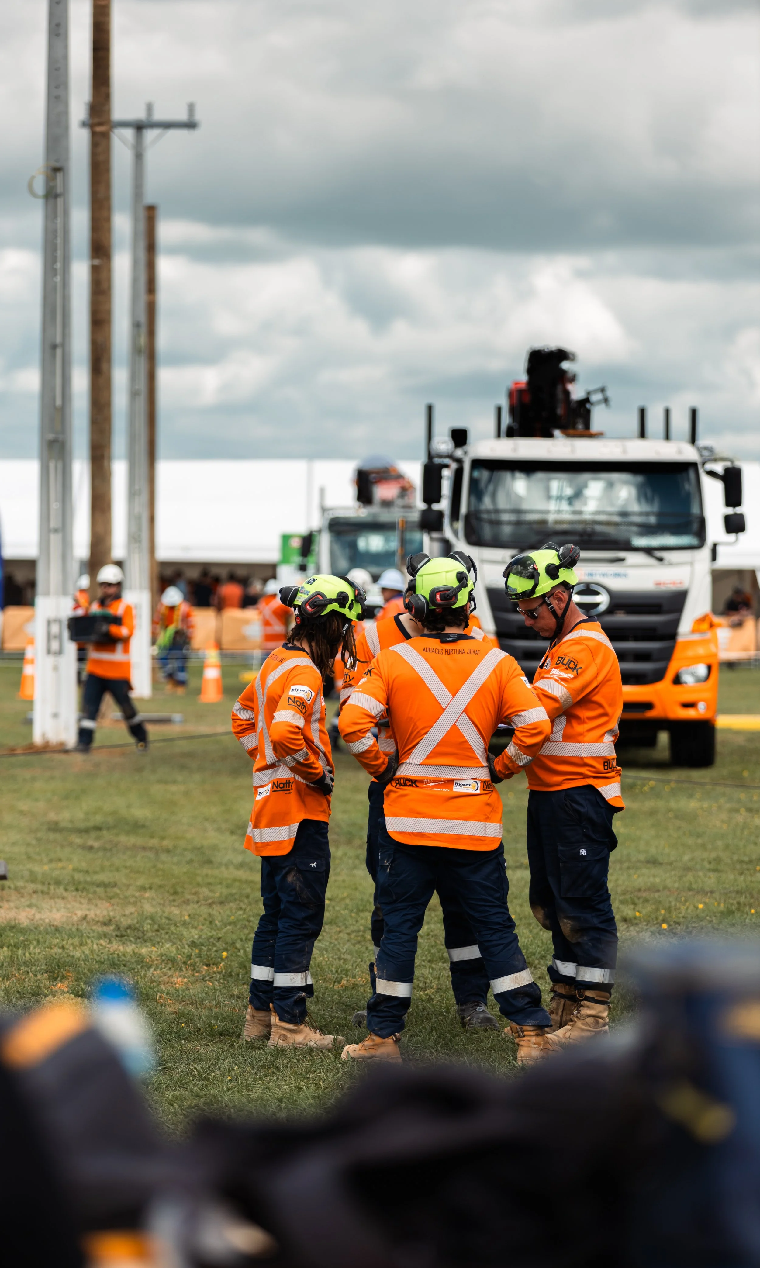Group of construction workers in orange safety vests and helmets standing in a circle on a grassy field, with industrial trucks and equipment in the background.