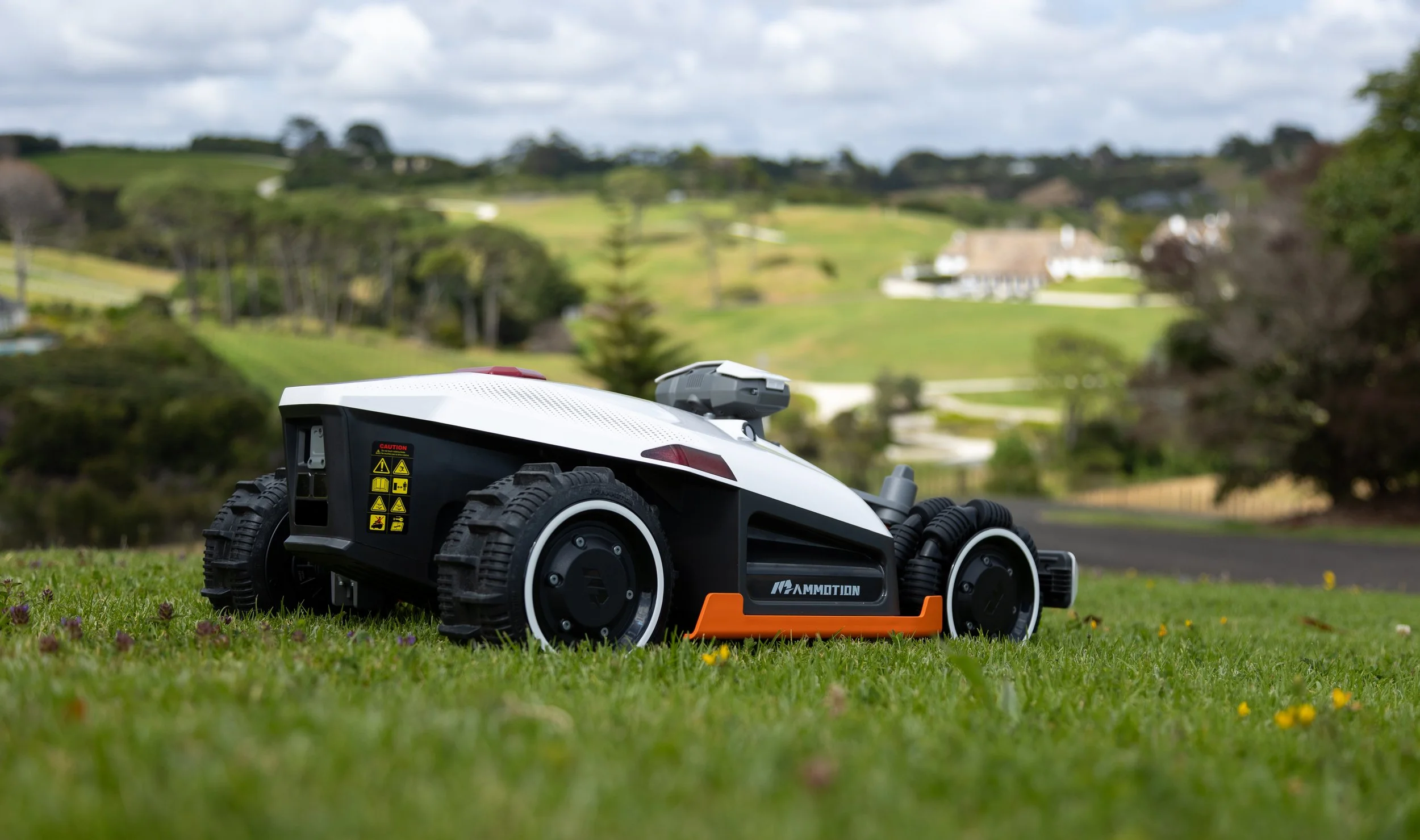 A robotic lawnmower operating on a grassy lawn with rolling hills and trees in the background.
