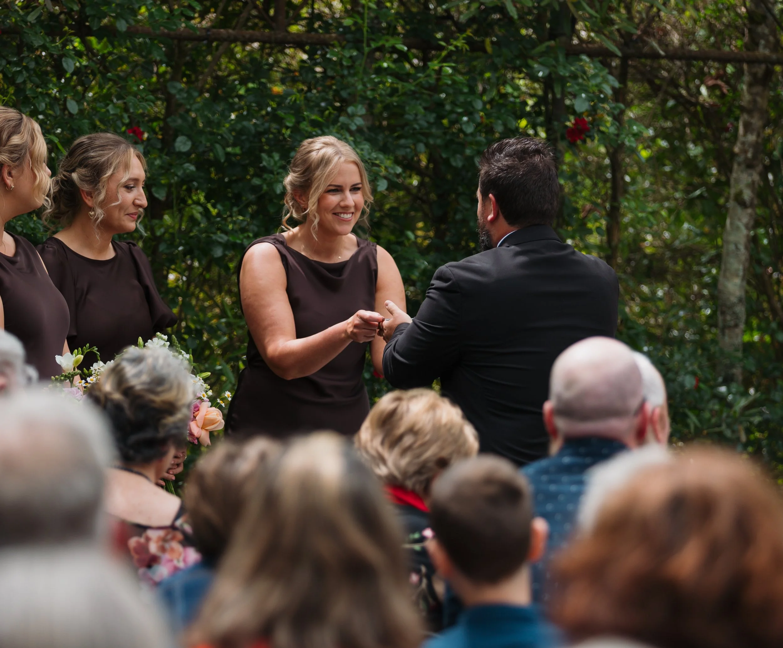 A woman with blonde hair in a dress holding a man's hand during a wedding ceremony outdoors, surrounded by guests and greenery.