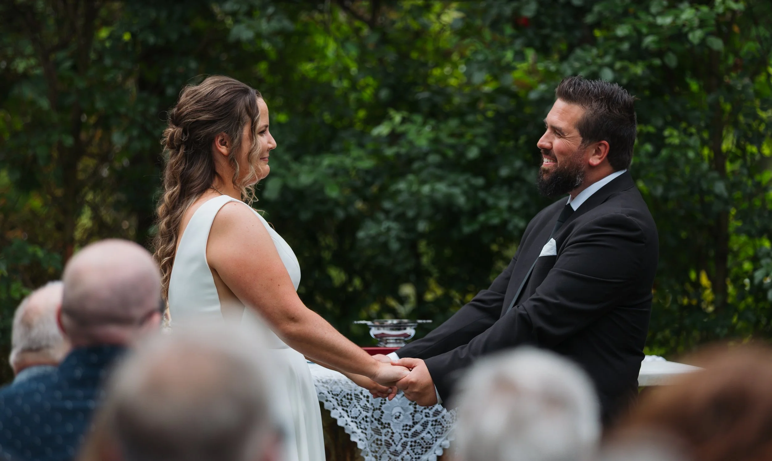 A bride and groom holding hands and smiling at each other during an outdoor wedding ceremony, with guests in the foreground and lush green trees in the background.