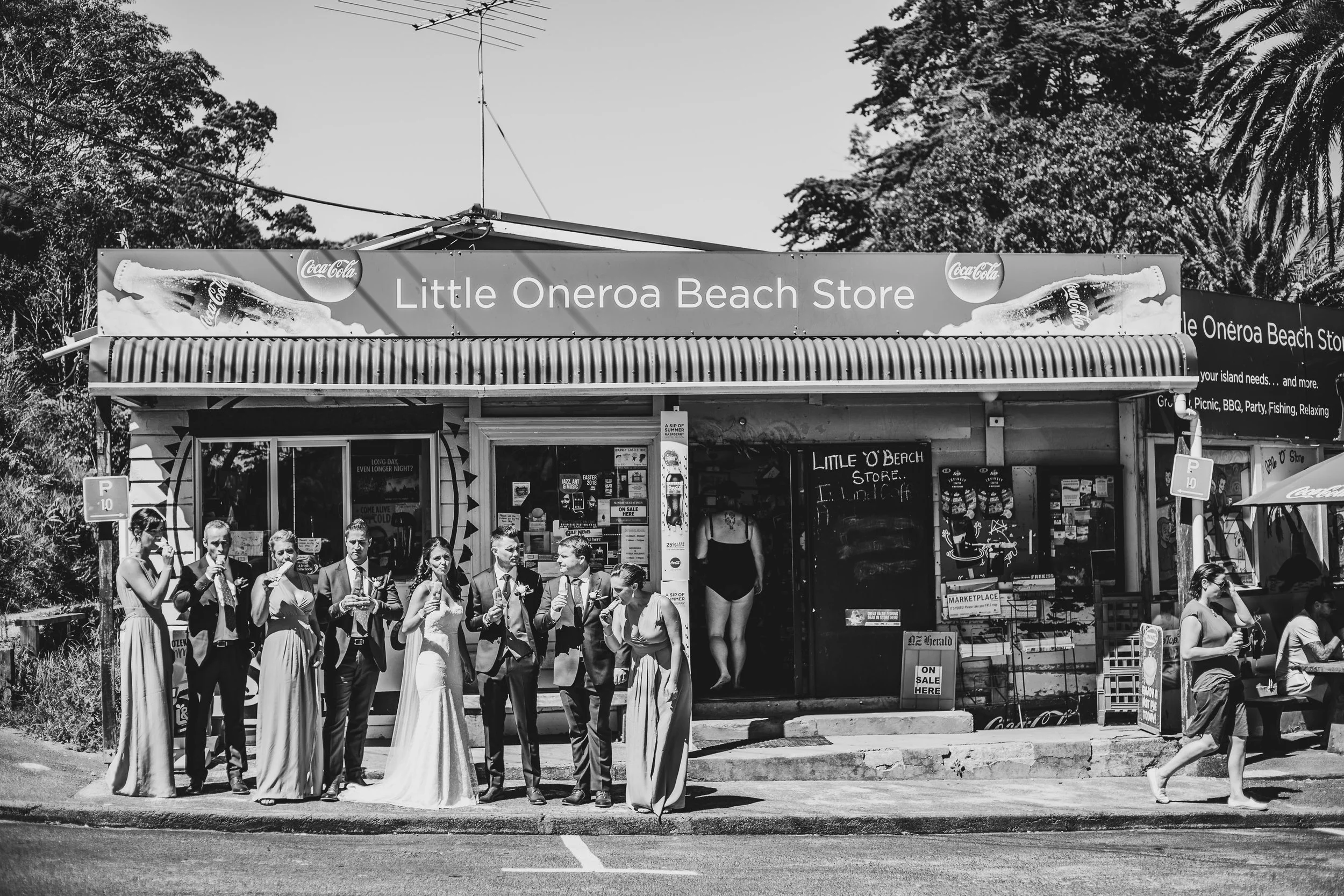 Group of people in wedding attire standing outside Little Oneroa Beach Store with a surfboard advertisement on top
