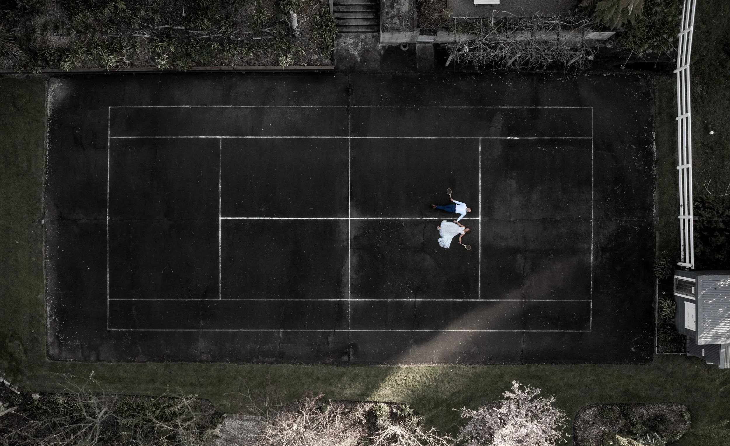 An aerial view of a dark outdoor tennis court with two people dressed in wedding attire, holding tennis rackets, lying on the court near each other. Old Forest School wedding venue