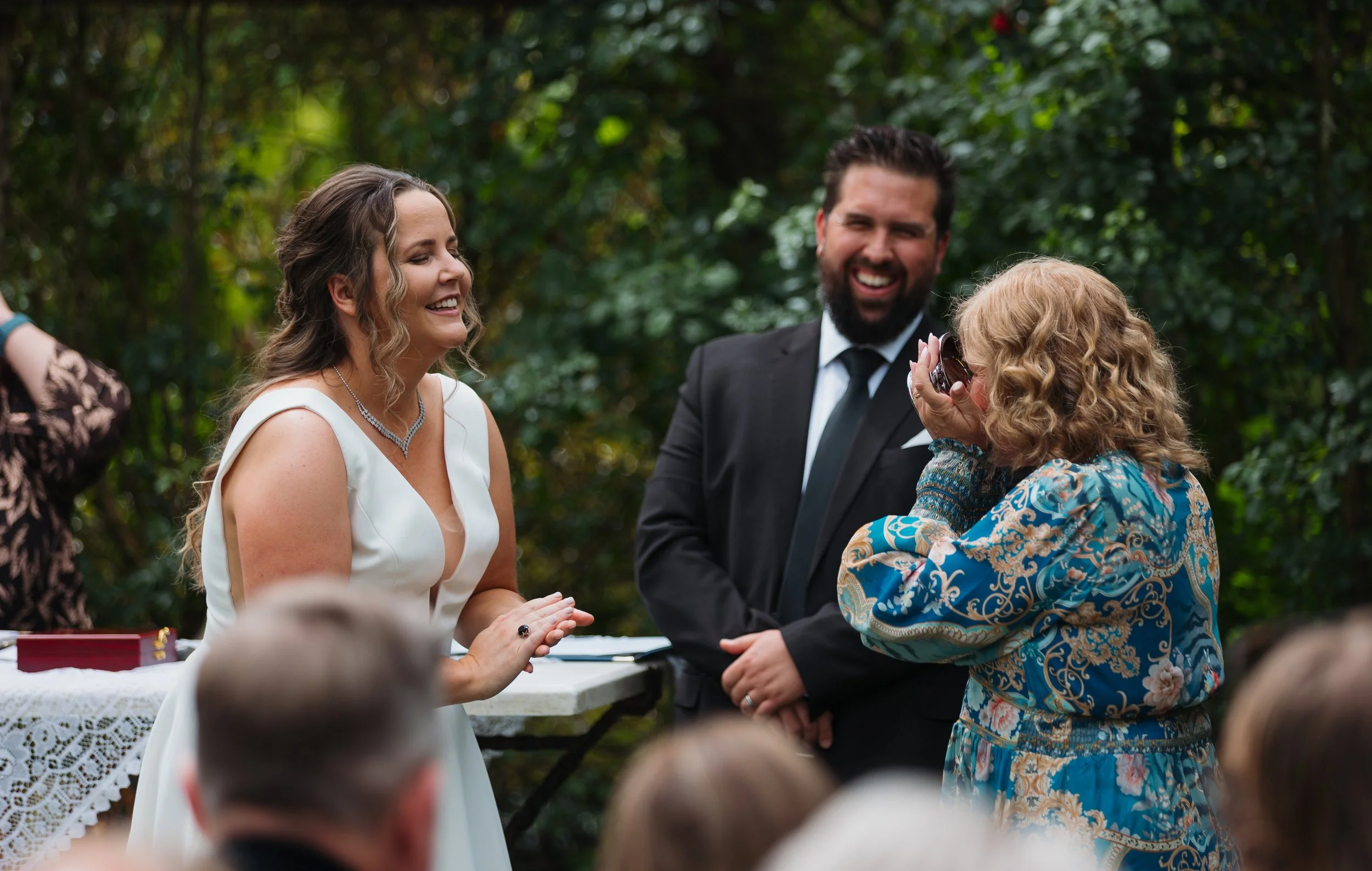 Bride in white dress smiling and gesturing during her wedding ceremony outdoors, with a man in a suit and a woman in a colorful dress laughing nearby, surrounded by greenery.