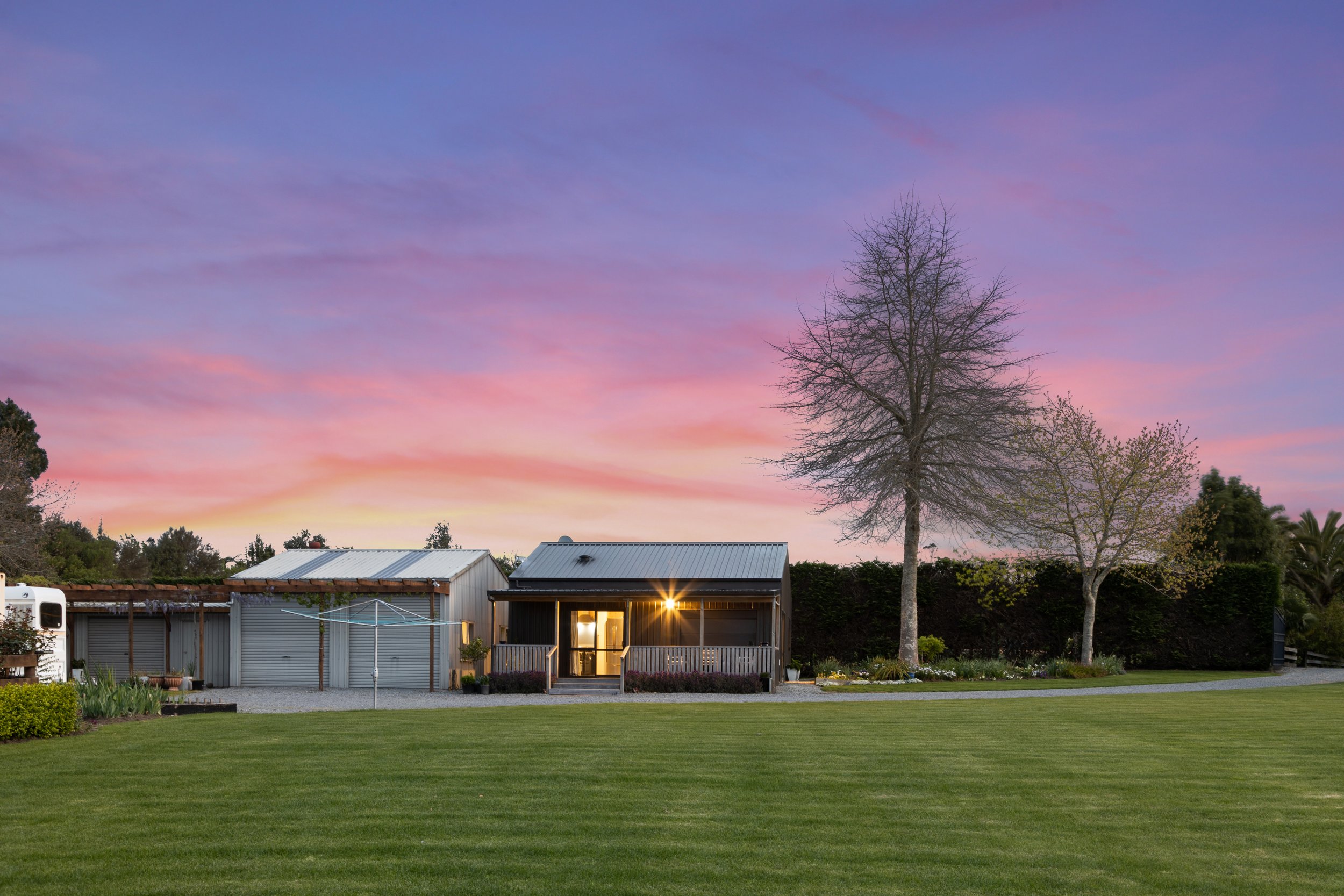 A house with a porch and a light on, surrounded by trees and a well-maintained lawn at sunset with a pink and purple sky.