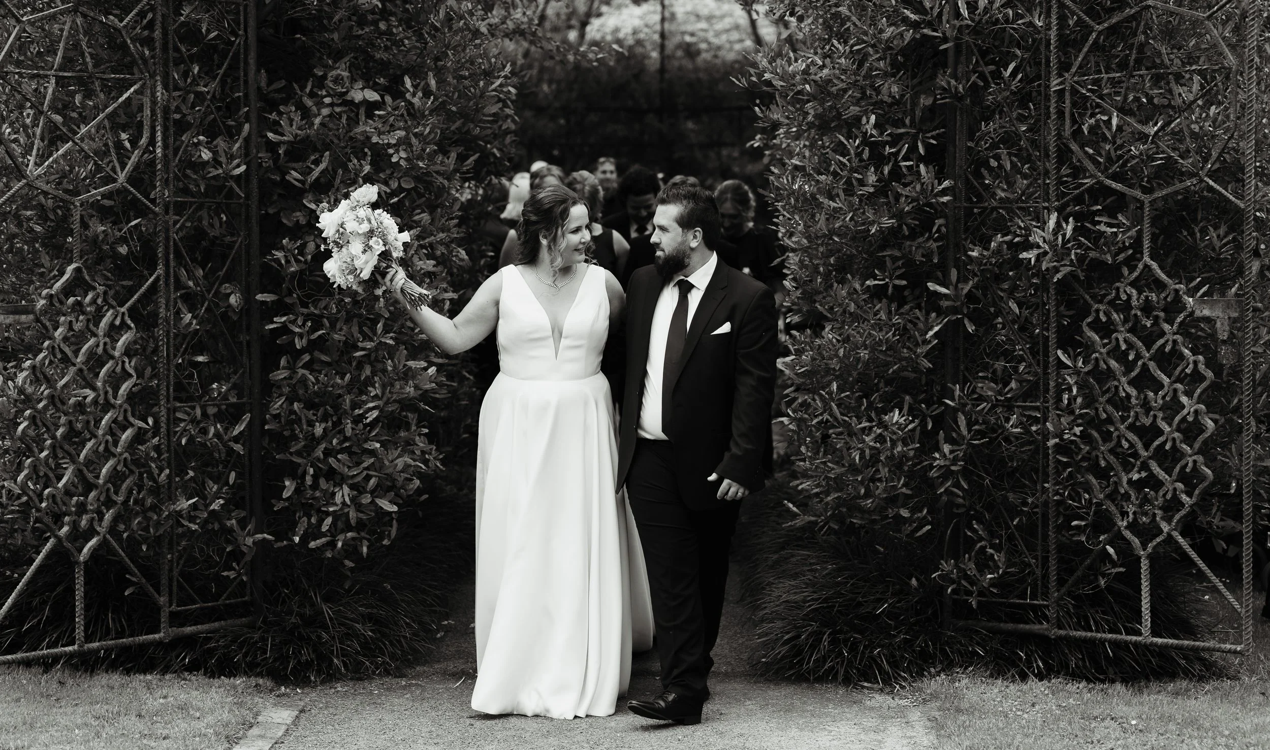 A newlywed couple walking through a garden archway, with the bride holding a bouquet and smiling at the groom, surrounded by guests. Tree church wedding venue