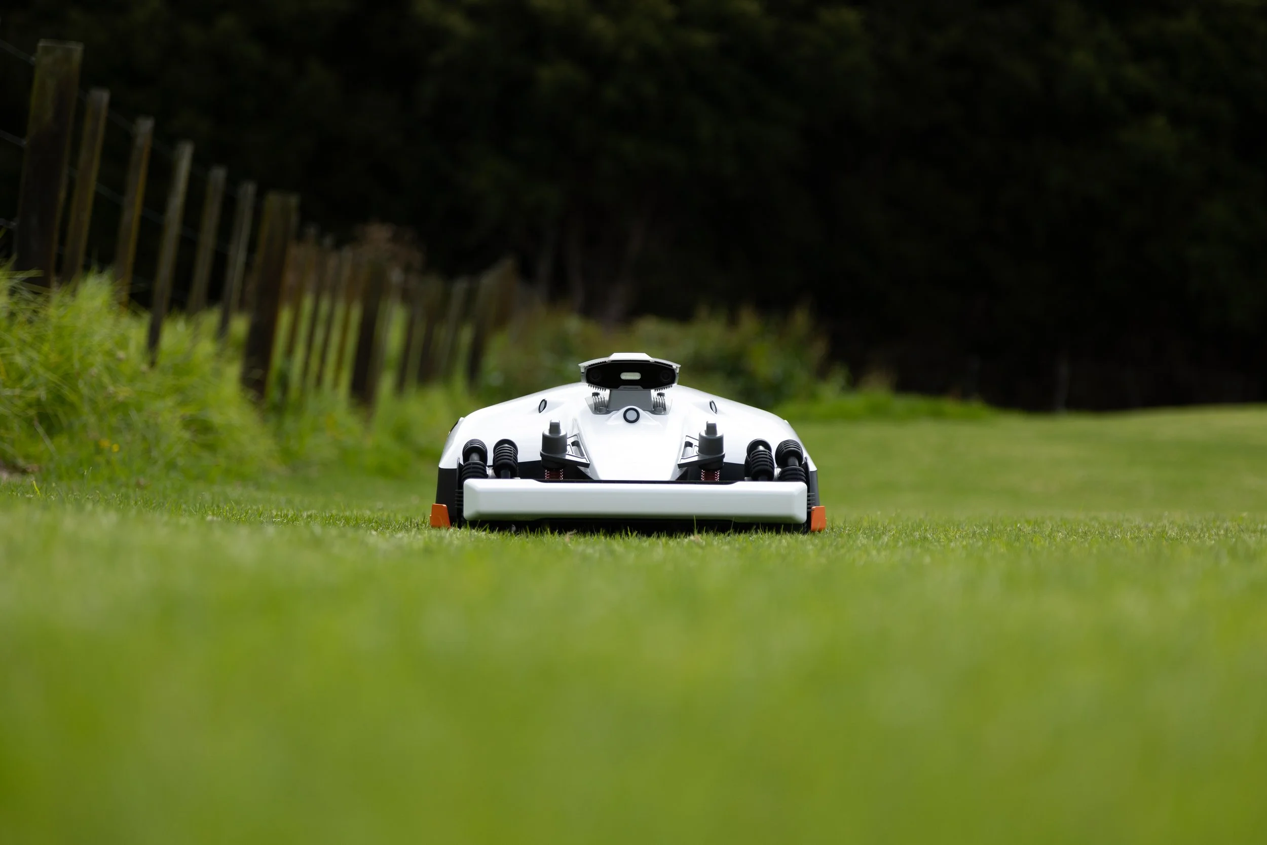 Autonomous robotic lawn mower on a grassy field with a fence and trees in the background. Robomate