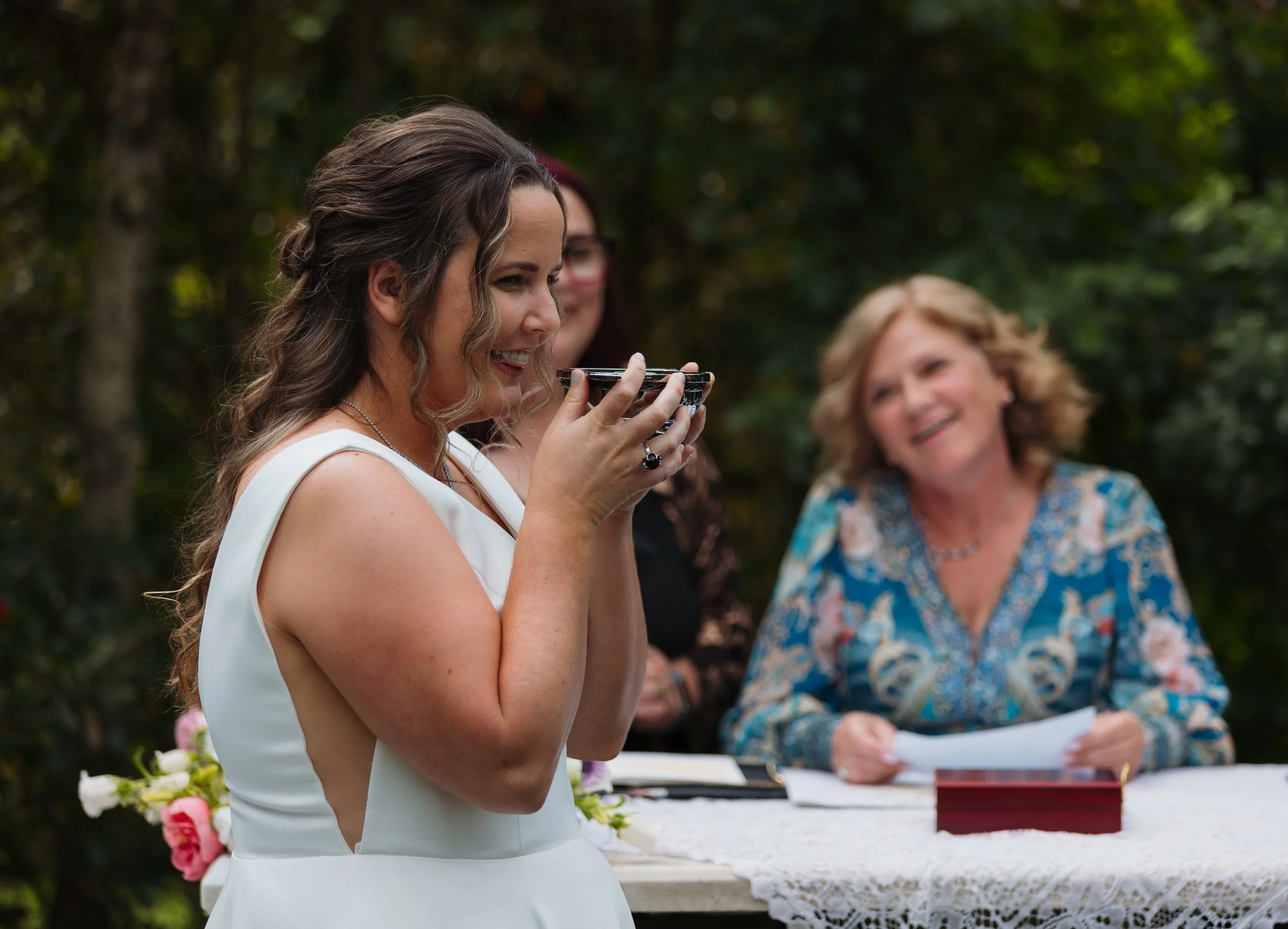 A woman in a white dress holds a small bowl close to her face during an outdoor wedding ceremony, with smiling women in the background.