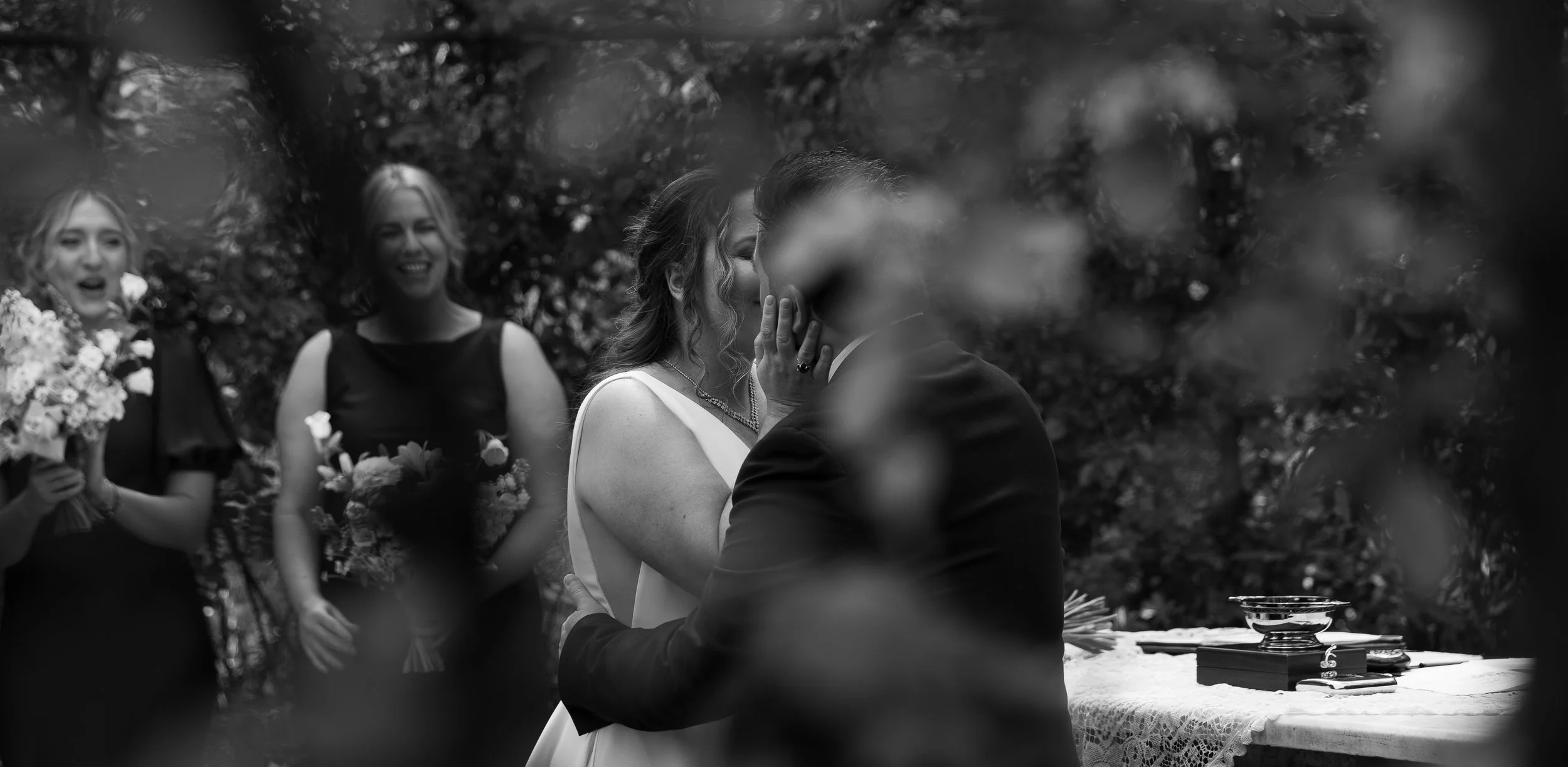 A black and white photo of a wedding ceremony, with a couple kissing at the altar surrounded by bridesmaids holding flowers, while trees are in the background.