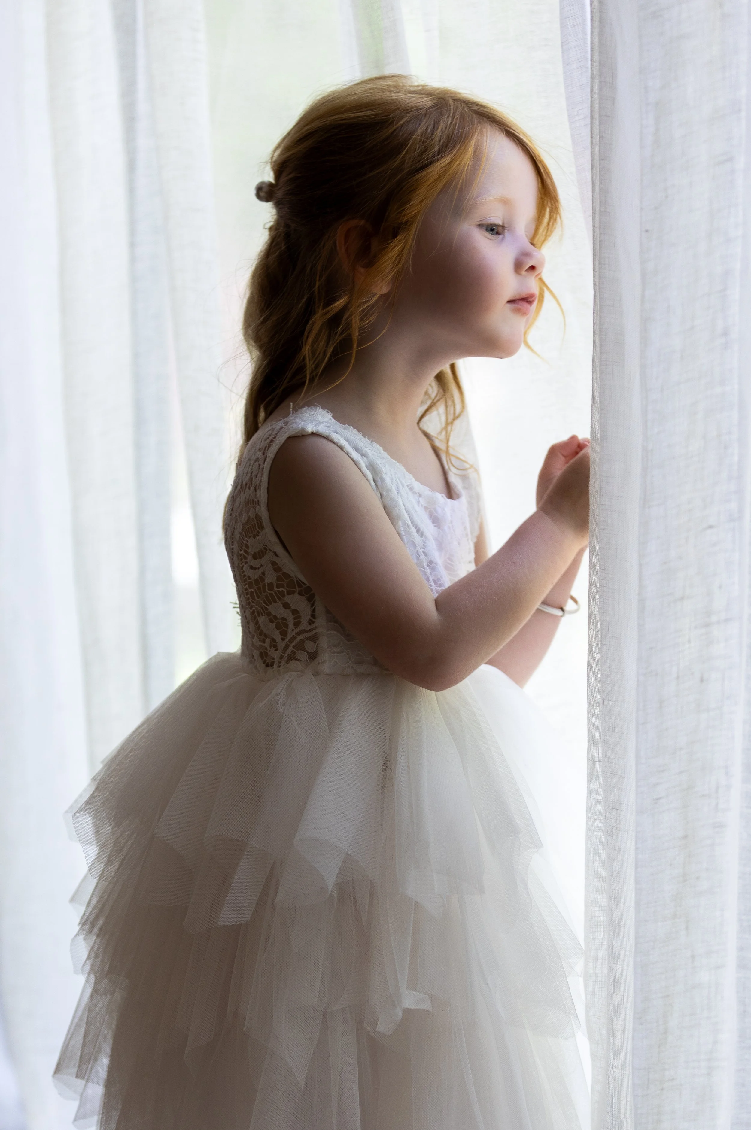 A young girl with red hair wearing a white lace dress and tulle skirt, standing beside a window with sheer curtains, looking outside thoughtfully.