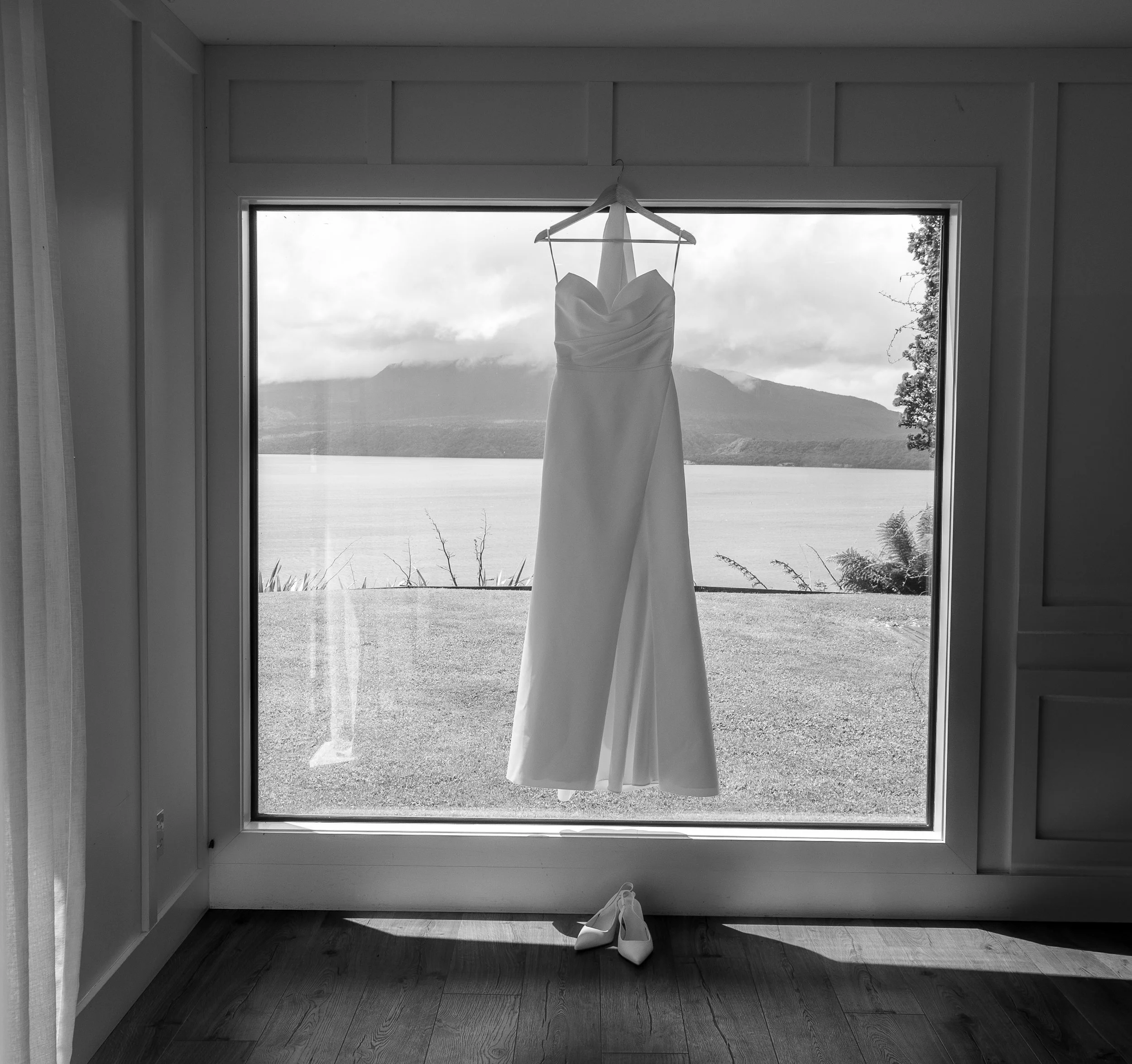 A wedding dress hanging on a hanger in front of a large window, with a scenic view of mountains, lake, and cloudy sky outside. A pair of shoes on the floor below the dress. Blackbarn wedding venue