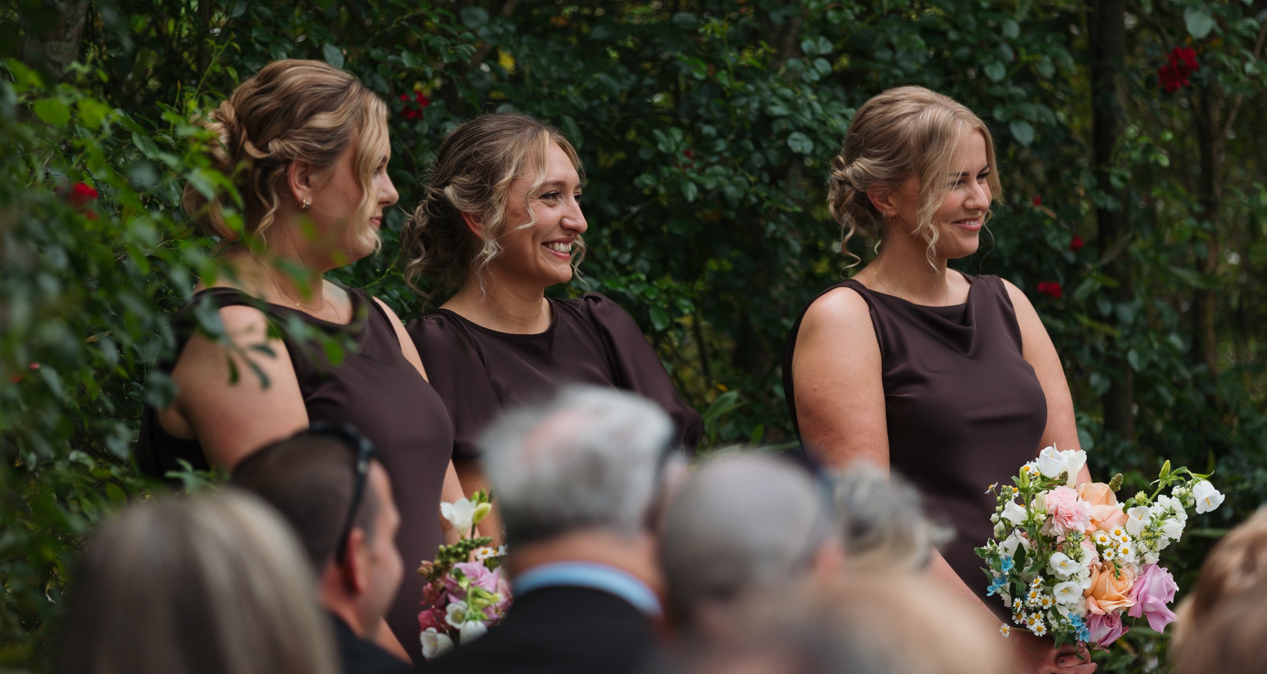 Three women in brown dresses smiling and holding bouquets of flowers at an outdoor gathering with green foliage in the background.