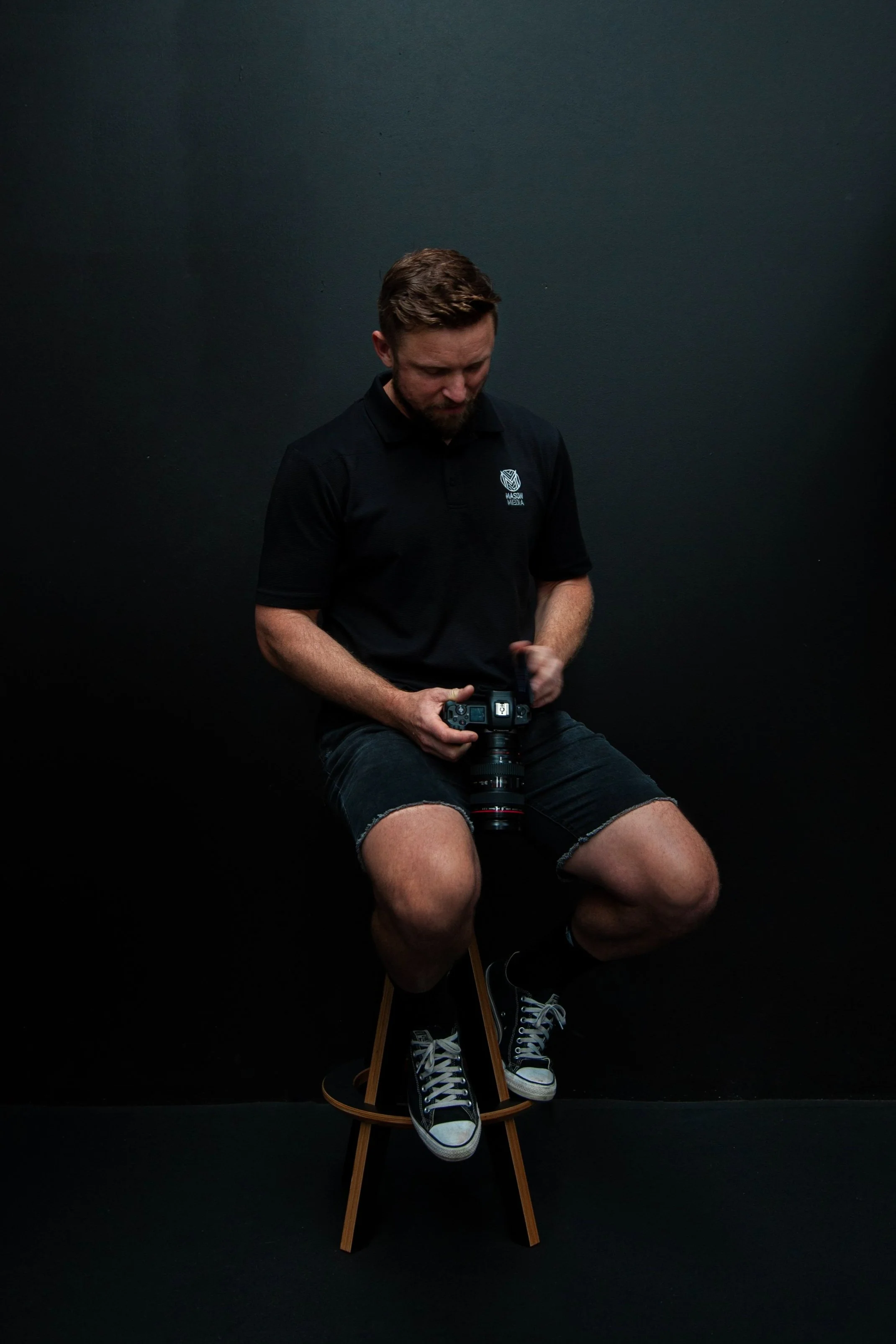 Young man with brown hair and beard, wearing a black polo shirt, black denim shorts, and black sneakers with white laces, sitting on a wooden stool against a black background, holding a camera in his hands.