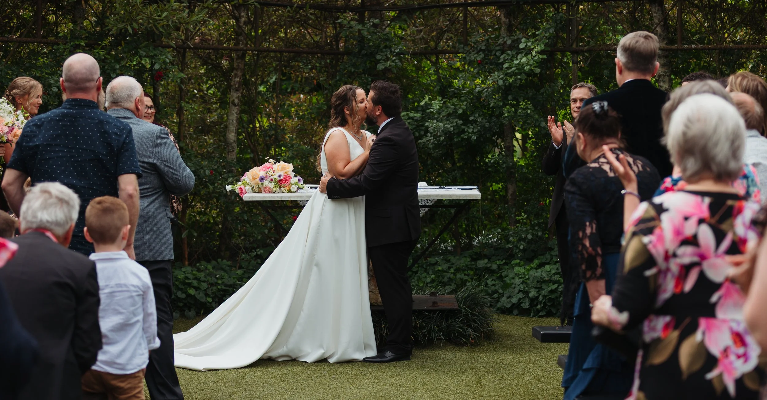 A bride and groom kiss during their outdoor wedding ceremony as friends and family watch and applaud.
