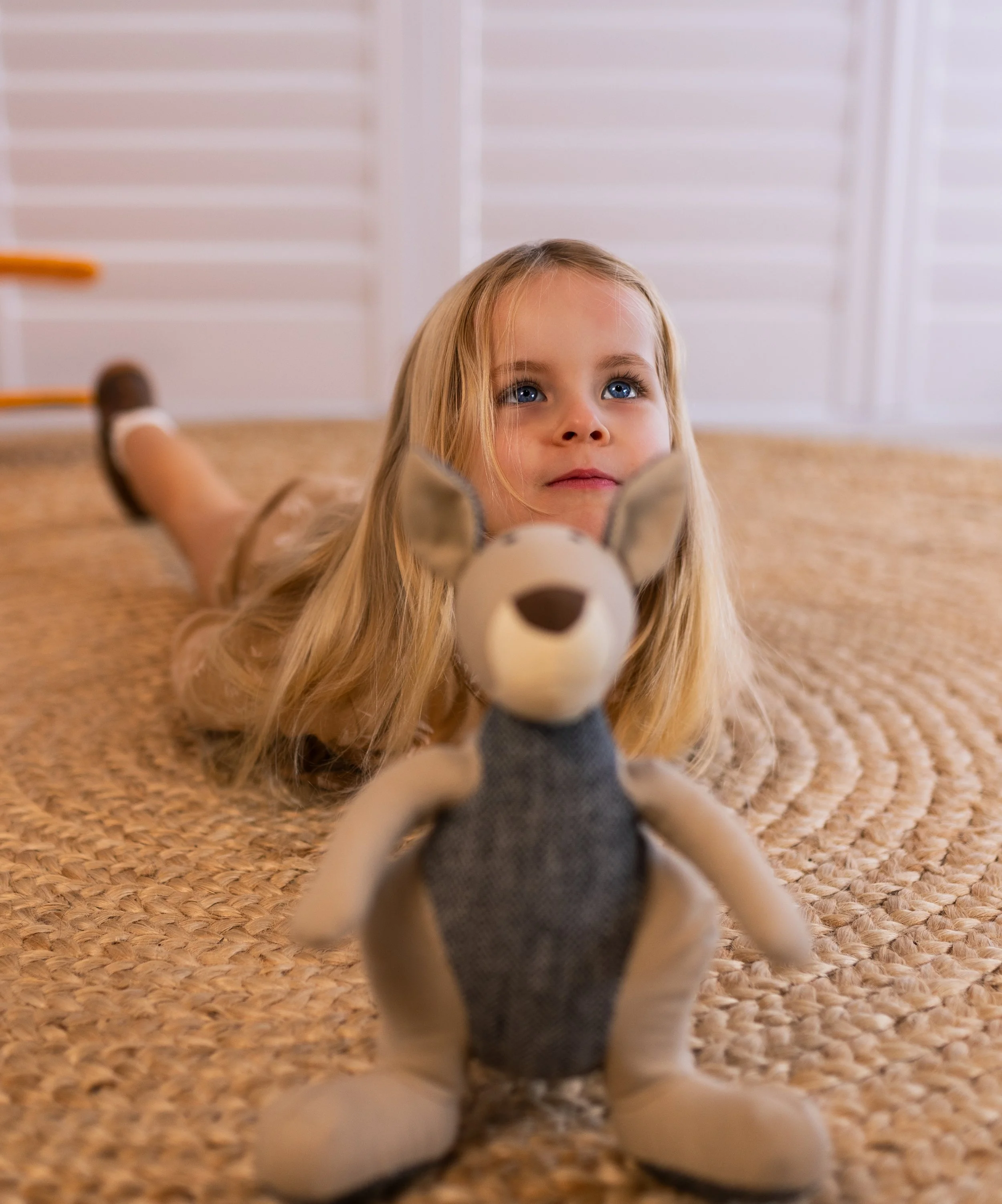 A young girl with long blonde hair and blue eyes lying on her stomach on a woven rug, holding a stuffed animal dog in front of her with a background of white window shutters.
