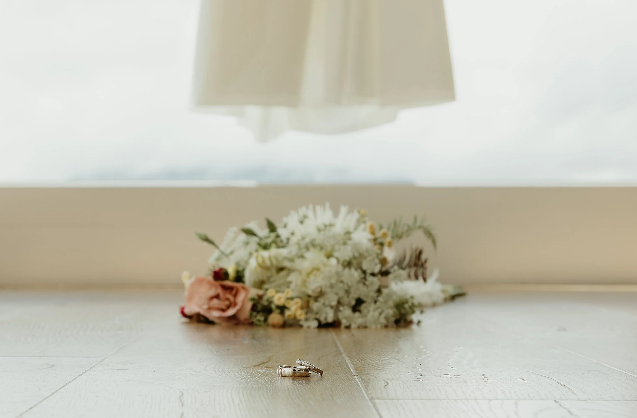 A wedding bouquet and two rings on a wooden floor with a large window in the background.