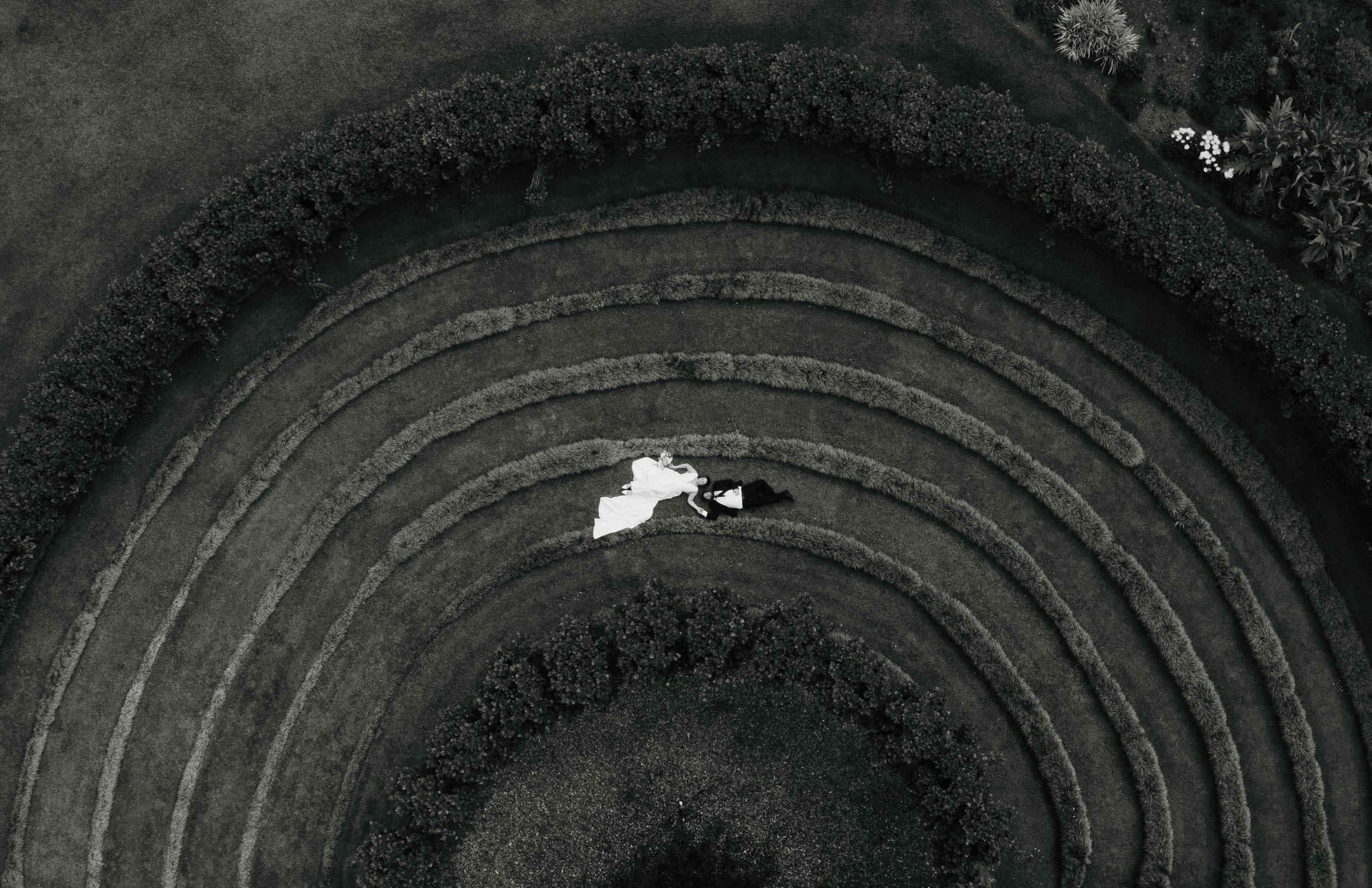 Black and white aerial photograph of a bride and groom lying on the grass, surrounded by concentric circular garden beds and bushes.