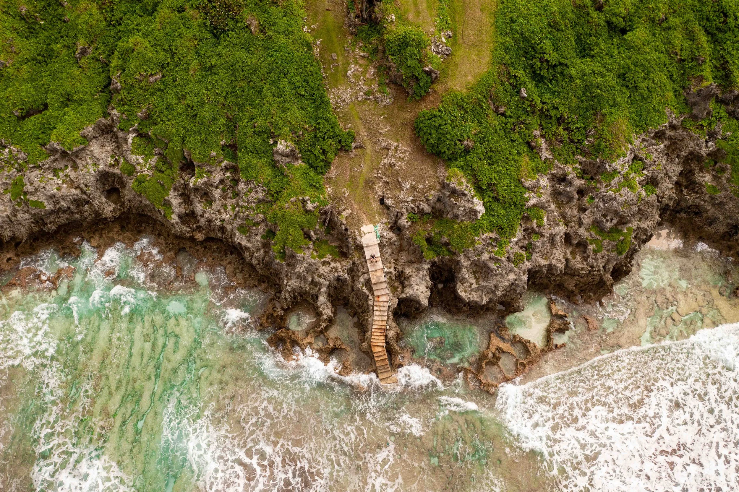 Aerial view of a rocky coastline with clear green water, a sandy beach, lush green vegetation, and a small wooden staircase leading to the beach. Niue