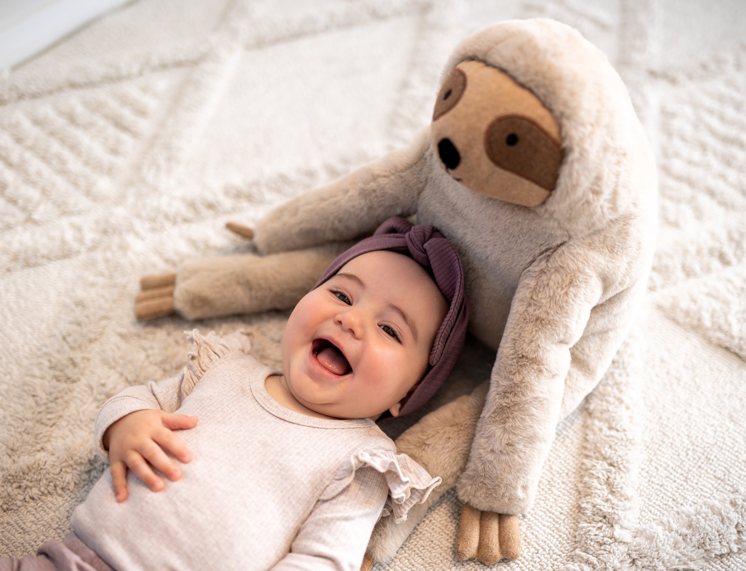 Smiling baby lying on a blanket with a stuffed sloth toy behind them.