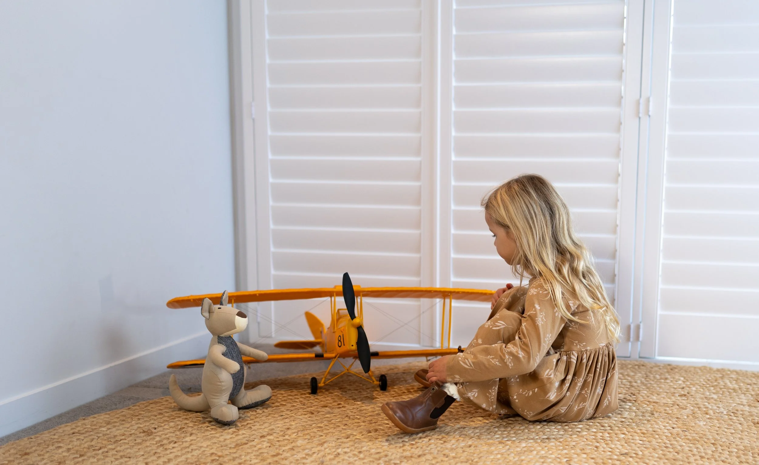 A young girl sitting on a woven rug, playing with a yellow toy airplane and a plush dog toy in a room with white plantation shutters.