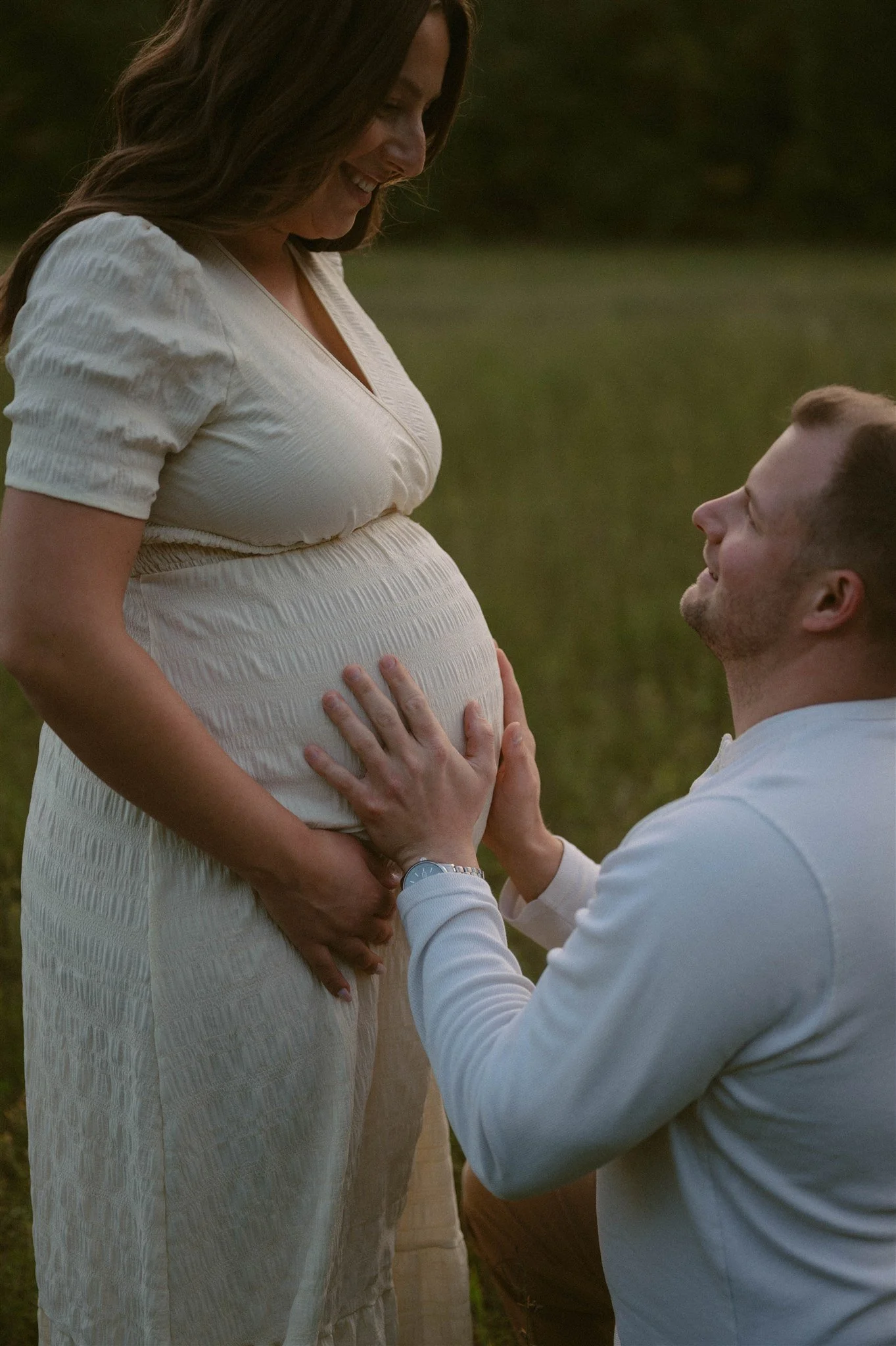 A pregnant woman in a white dress is smiling as a man kneels in front of her, holding her belly and looking up at her affectionately outdoors near a body of water.