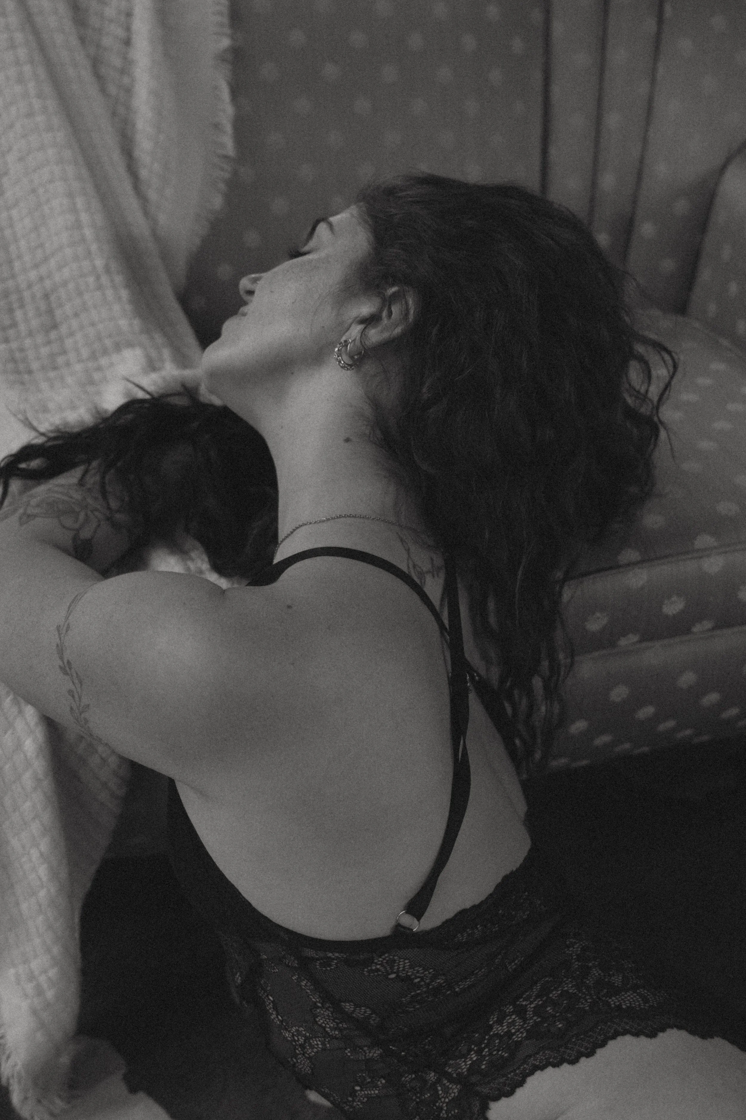 A woman with curly dark hair lies on a bed, wearing a lace camisole, with her eyes closed, in a black and white photo.