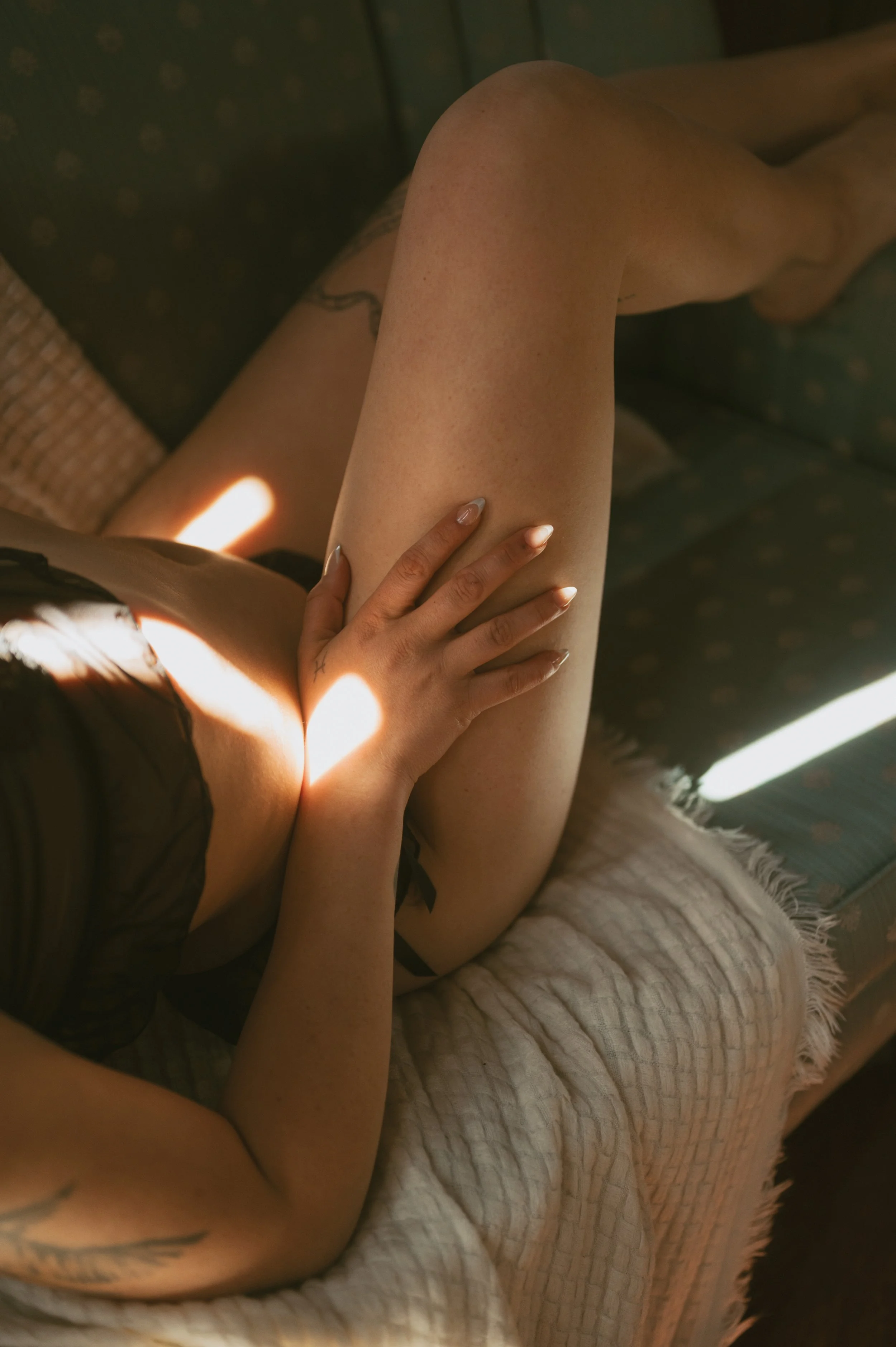 Close-up of a woman's legs with a hand resting on her thigh, illuminated by warm sunlight, with a sofa in the background.