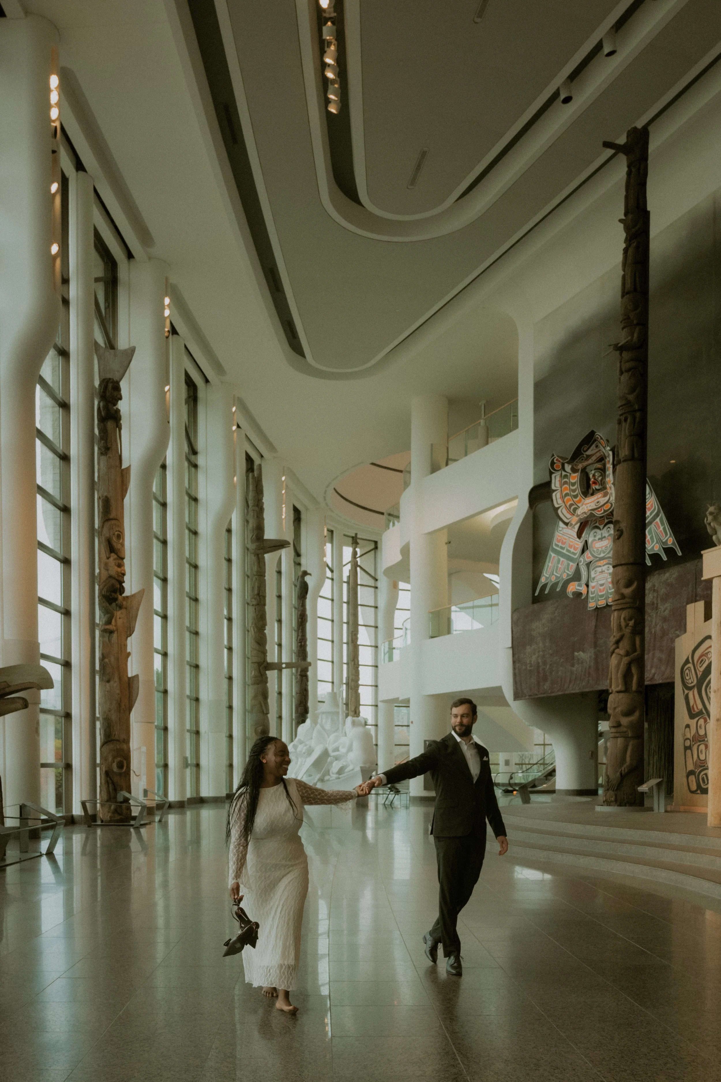 A bride and groom holding hands and dancing in a spacious, well-lit hall with large windows, traditional carved totem poles, and indigenous artwork.