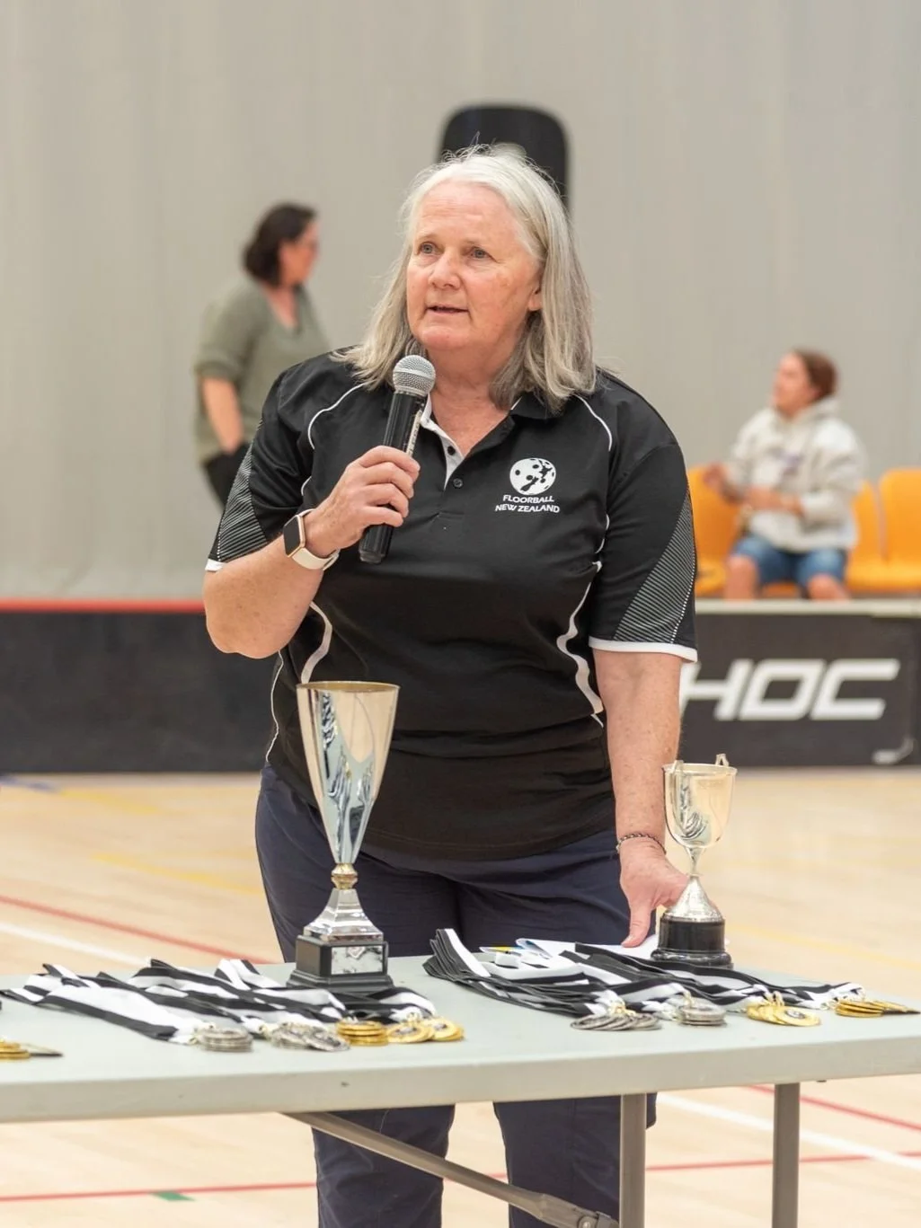 A woman with gray hair holding a microphone standing in front of a table with medals and trophies in an indoor sports facility.