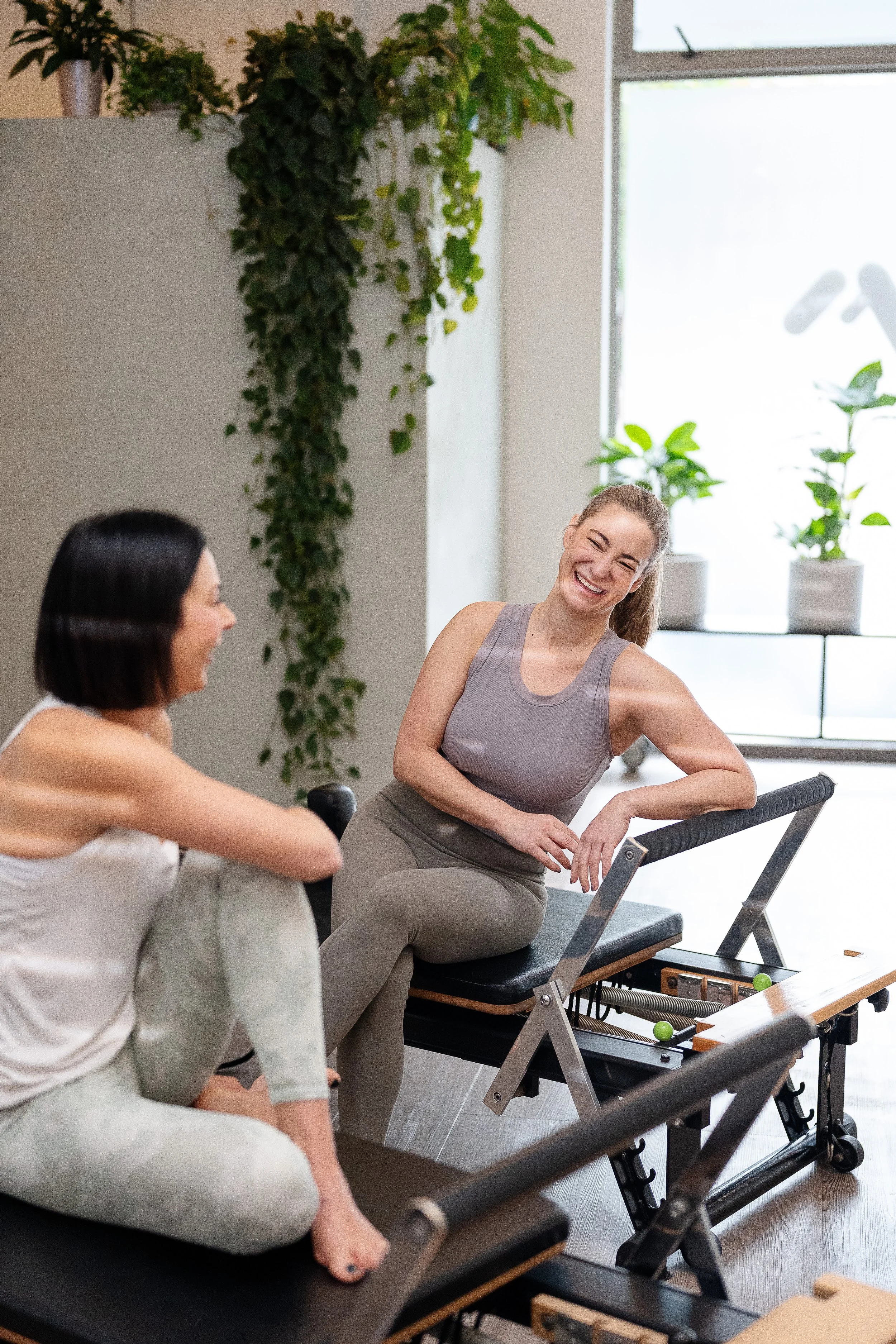 Two women seated on Pilates reformer machines in a bright, modern fitness studio, smiling and enjoying their workout. There are potted plants on a window sill in the background.