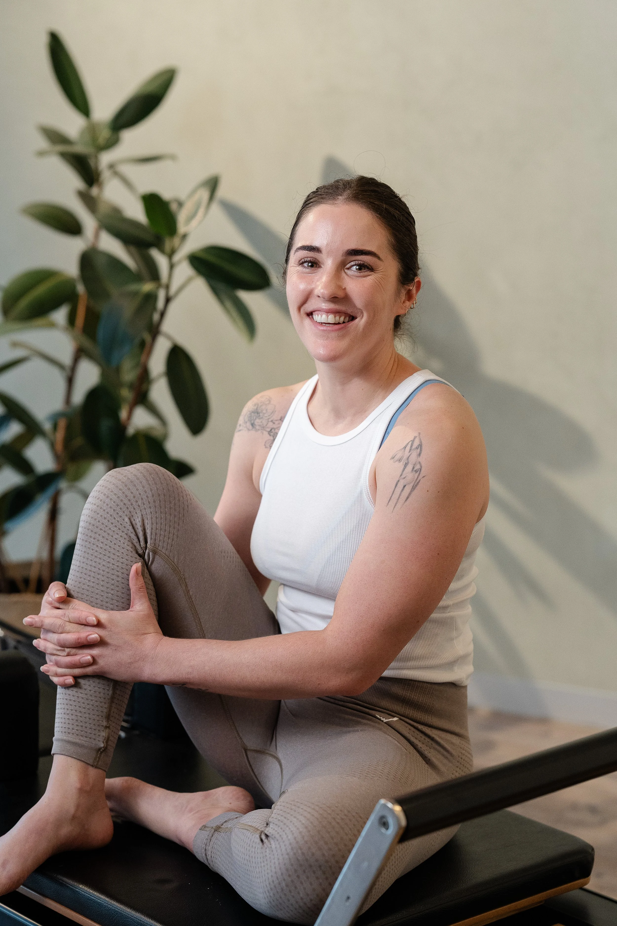 A woman smiling while stretching her leg on a Pilates reformer machine in a fitness studio, with a potted plant in the background.