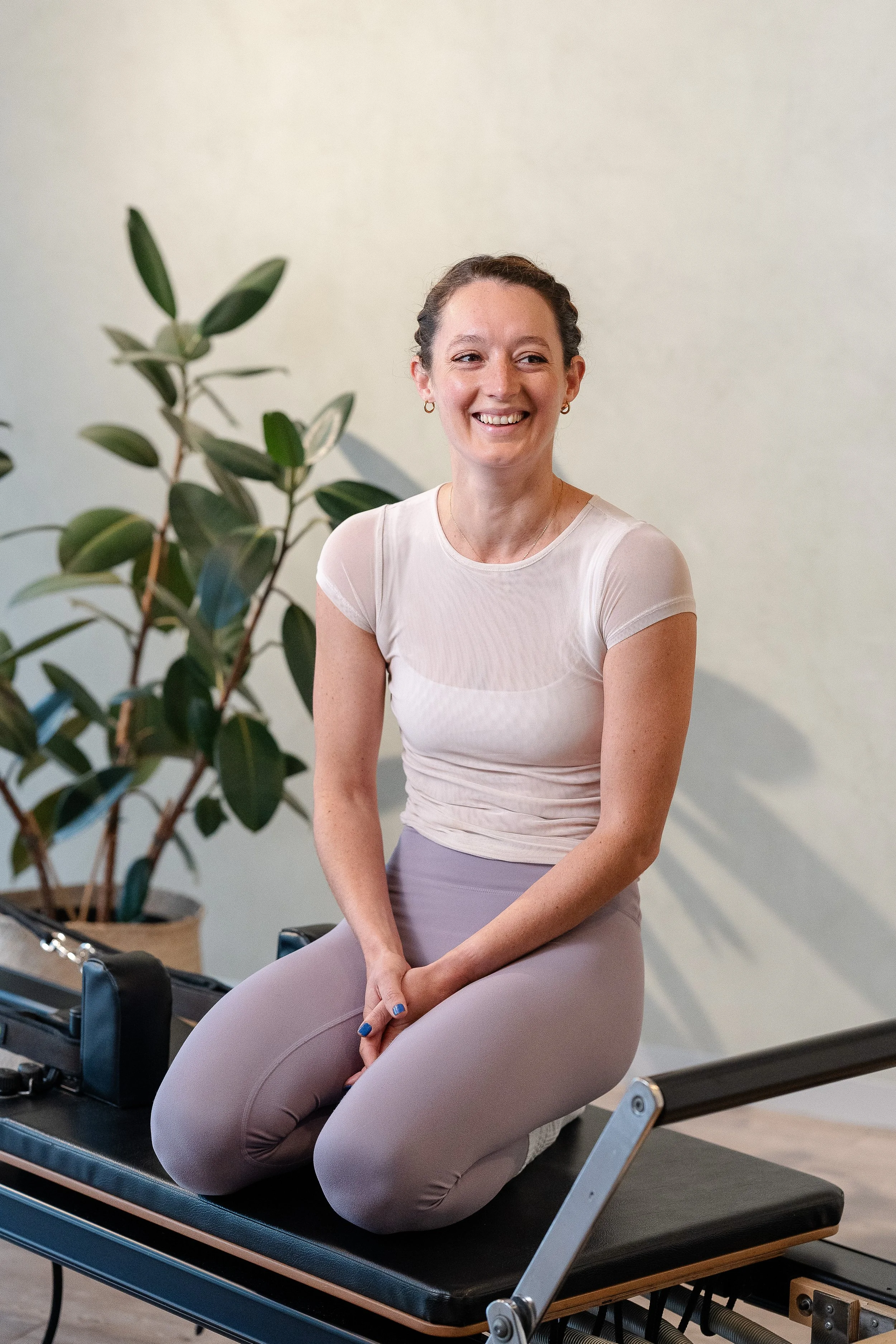 A woman smiling and sitting on a Pilates reformer machine in a fitness studio, with a potted plant in the background.
