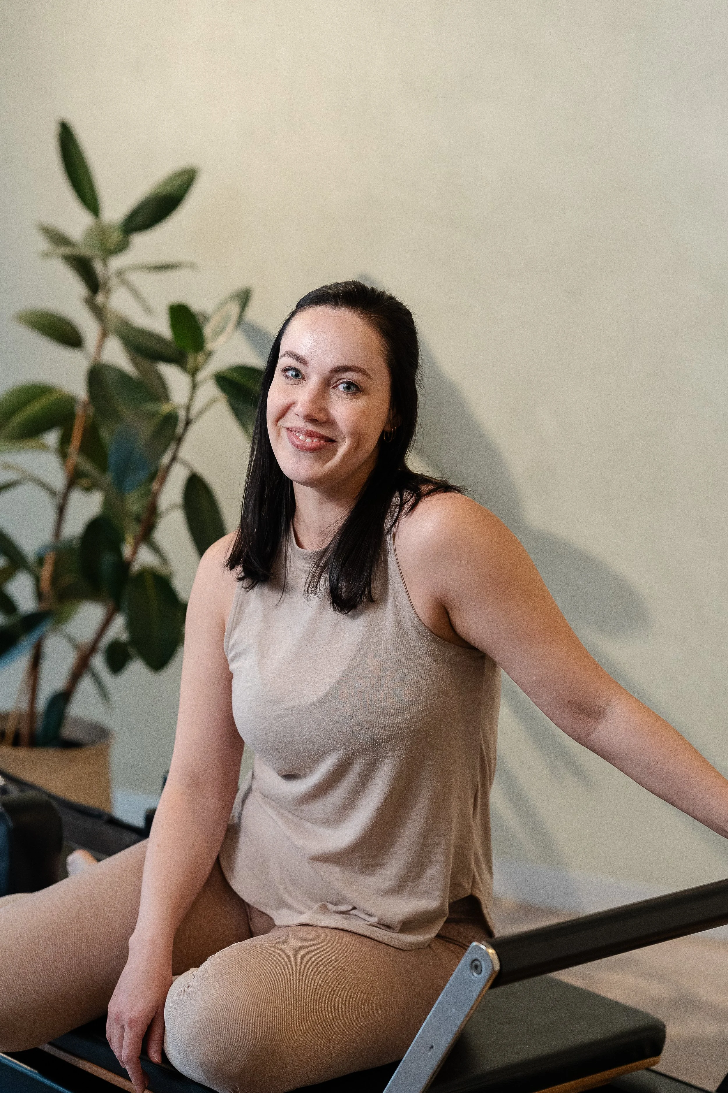 A woman with long dark hair, wearing a beige sleeveless top and matching beige pants, smiling and sitting on areformer chair in front of a light green wall and a plant with dark green leaves.
