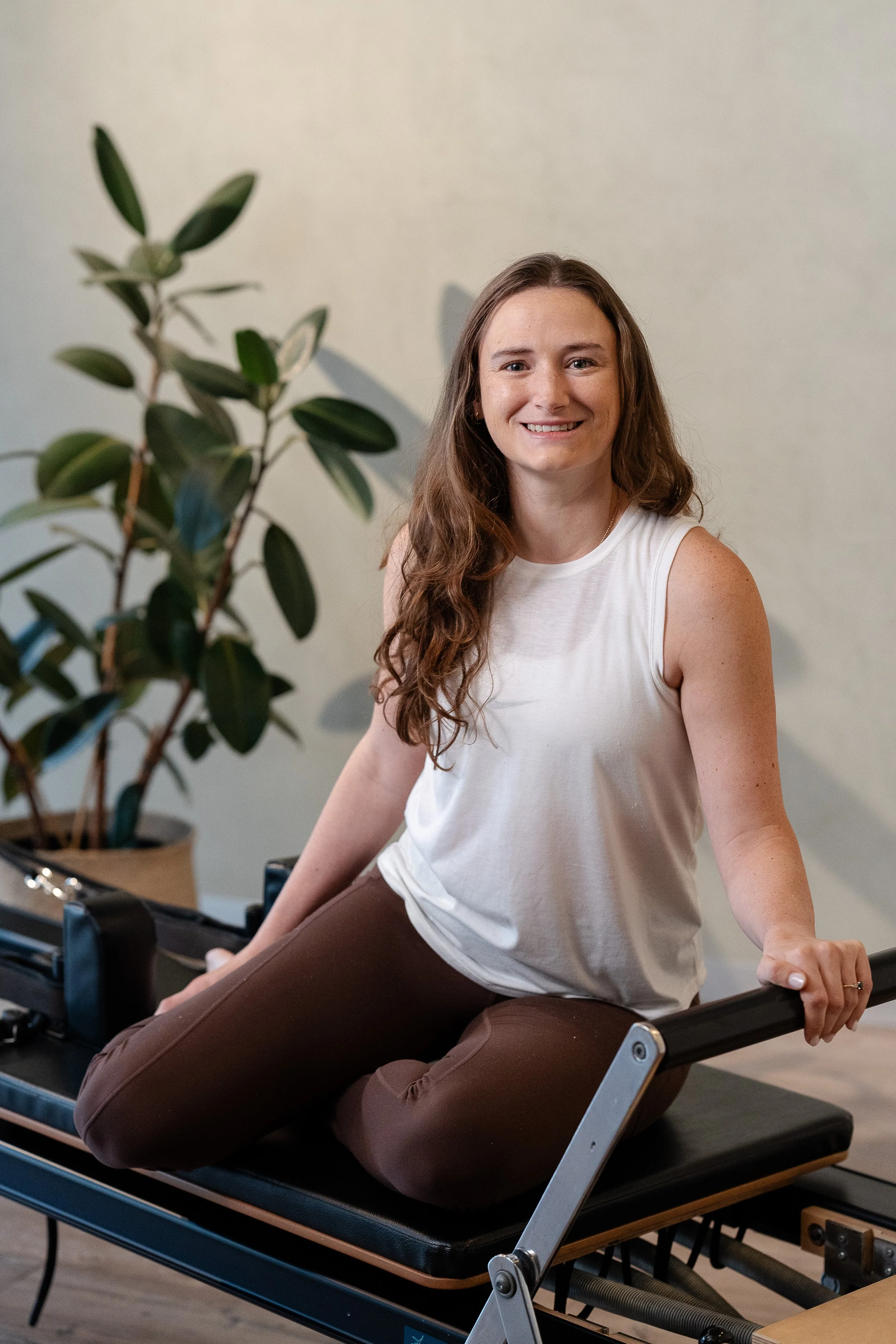 A young woman with long wavy brown hair, smiling, wearing a sleeveless white top and brown pants, sitting on a Pilates reformer machine in a fitness studio with a beige wall and a large green leafy plant nearby.