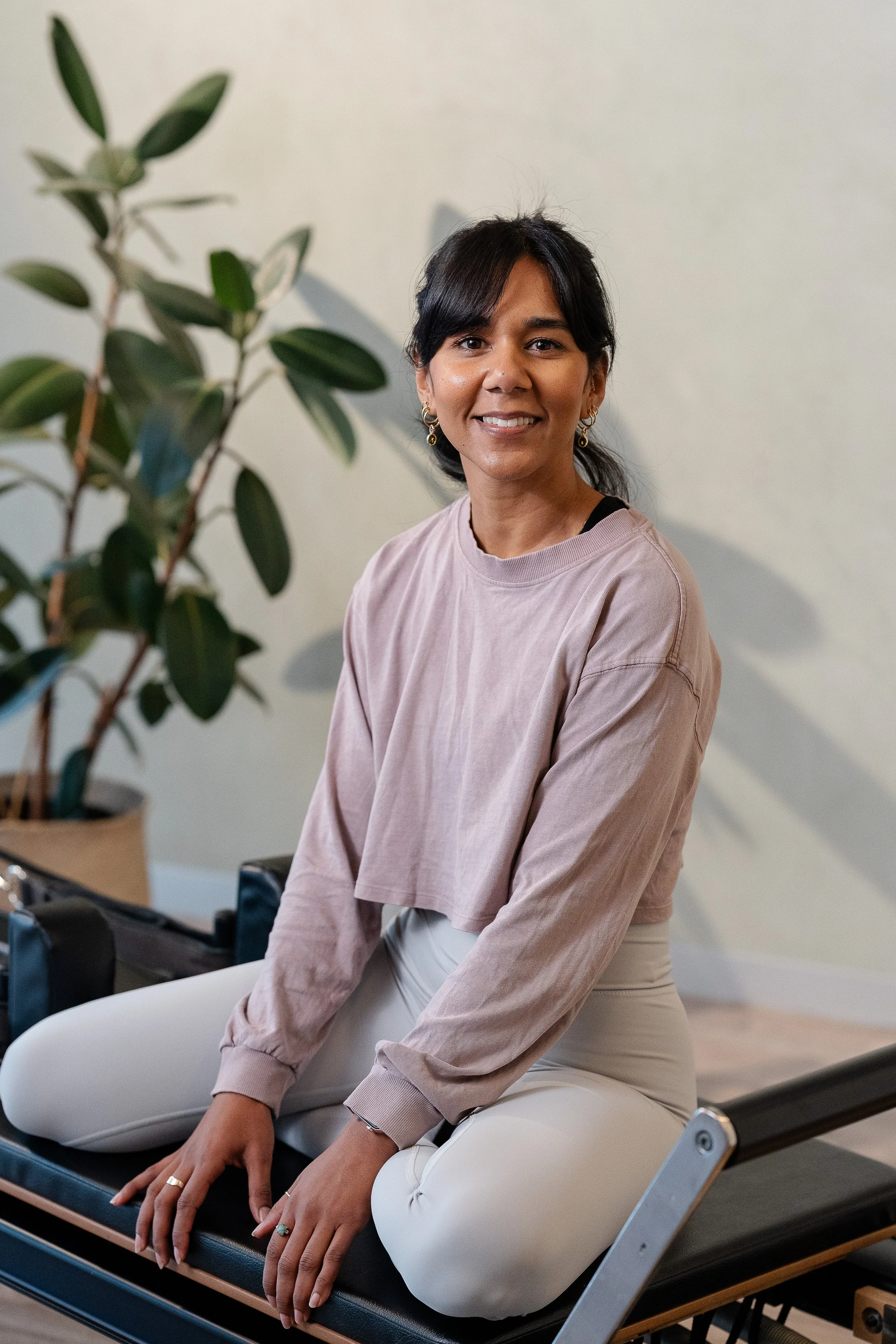 A woman with black hair wearing a light pink long-sleeve shirt and white pants sitting on a reformer, smiling at the camera. There is a large potted plant with green leaves to her left and a plain wall behind her.