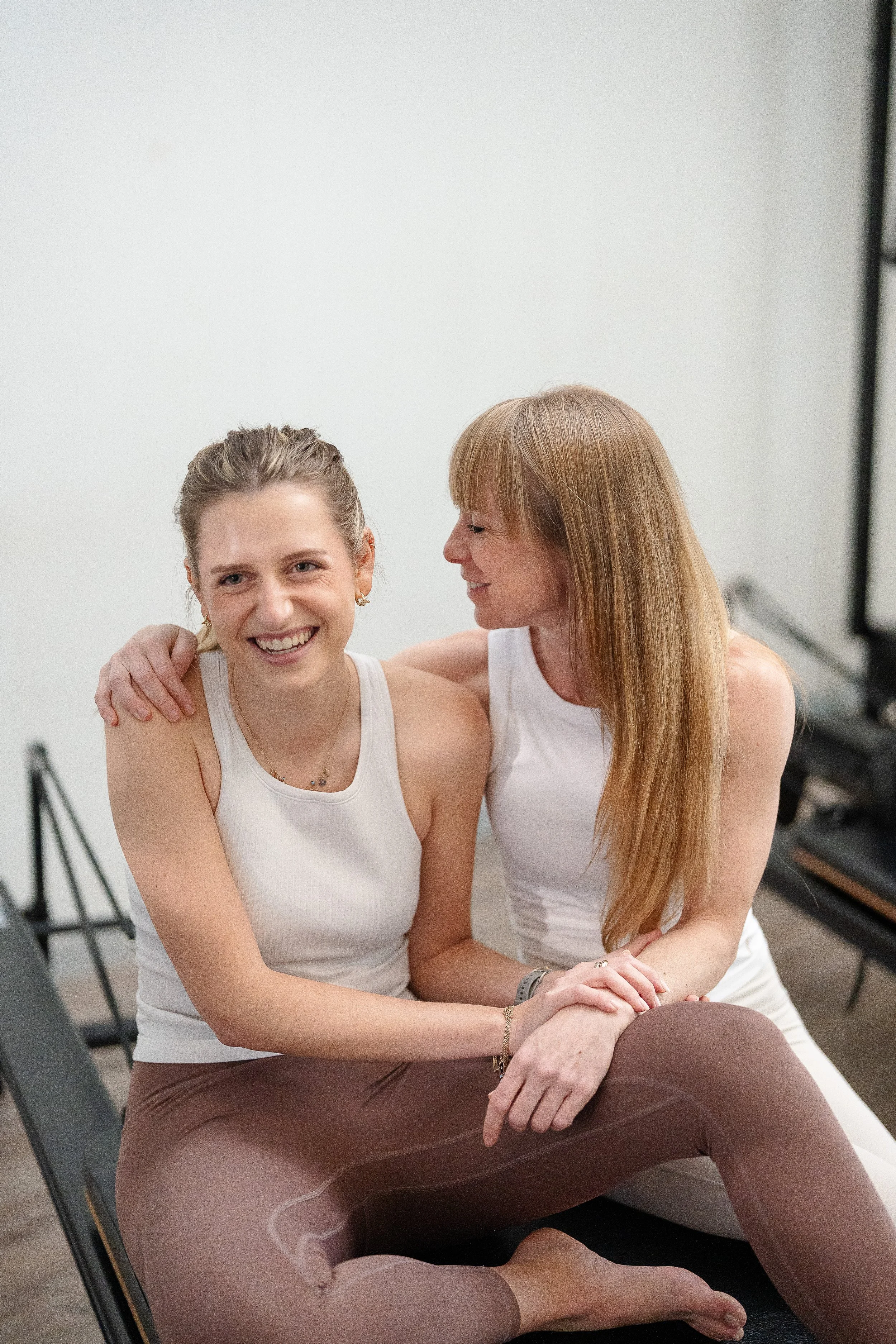 Two women sitting close together, engaged in conversation and smiling, in a bright indoor space. One woman has her arm around the other.