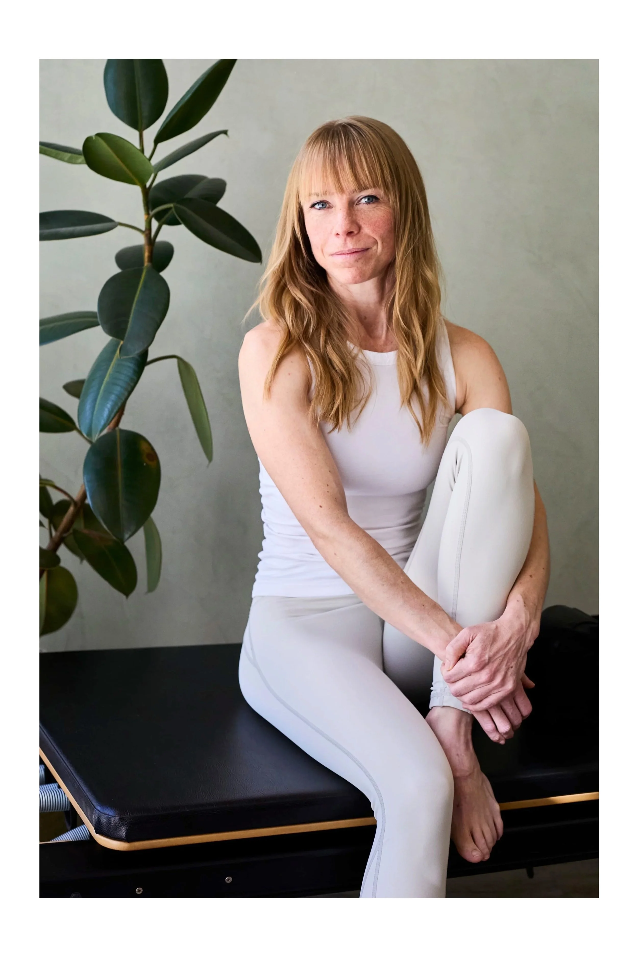 A woman with long red hair, wearing a white tank top and white leggings, sitting on a black examination table, holding her left foot with her right hand, next to a green plant with large leaves and a plain light grey wall.