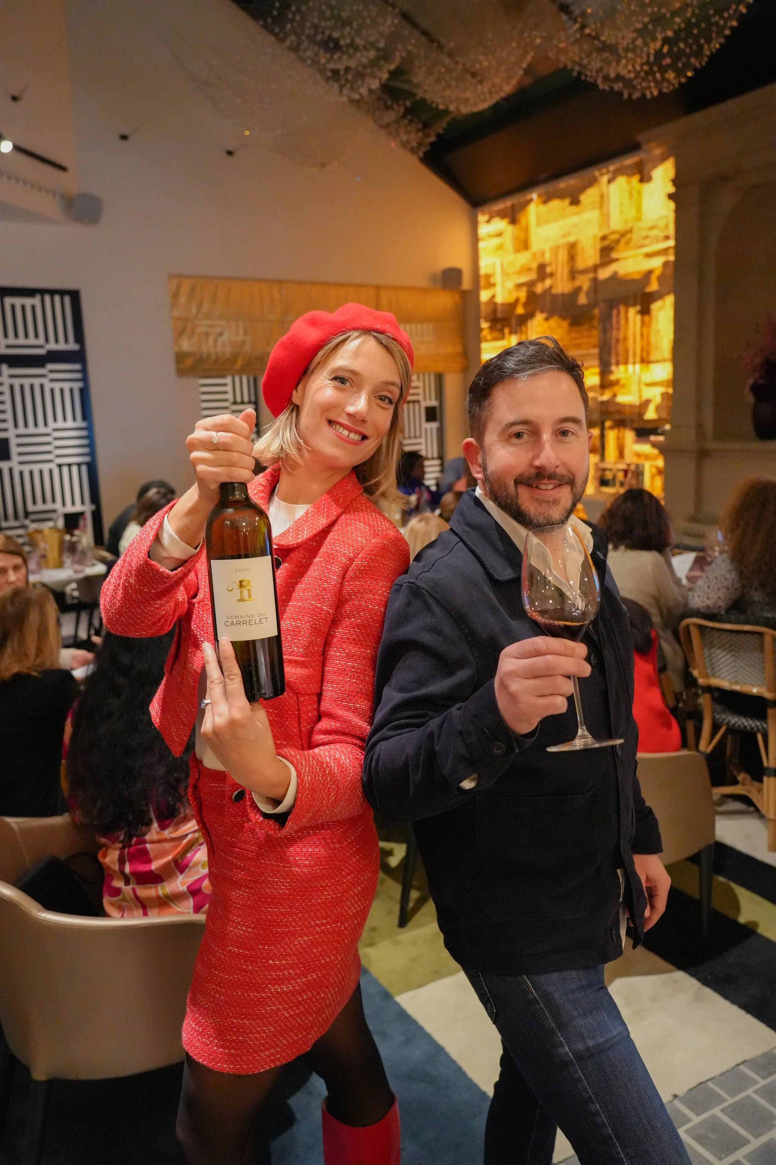 Deux personnes souriantes dans un restaurant, une femme en rouge avec un verre de vin, un homme en noir avec une bouteille de vin.