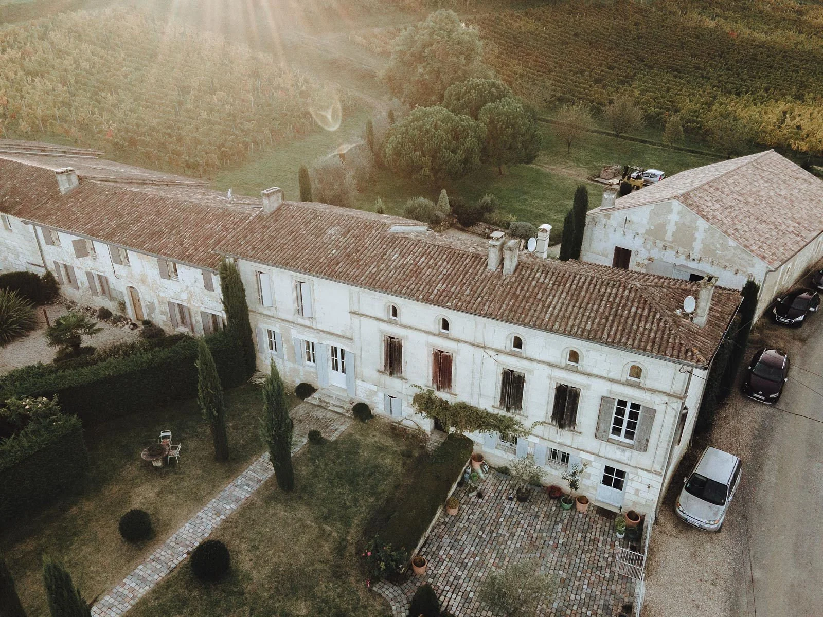 Vue aérienne d'une maison en pierre avec un toit en tuiles, entourée d'un jardin et de parking, avec un paysage rural en arrière-plan.
