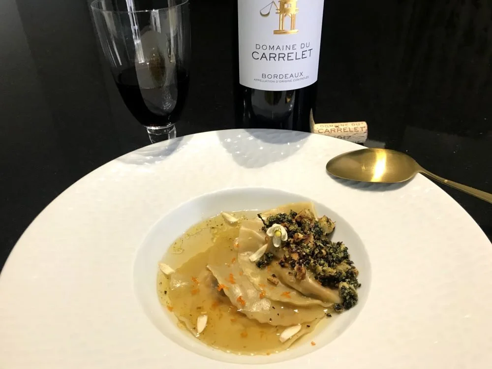 Une assiette de raviolis avec une garniture sombre et une sauce claire, accompagnée d'un verre de vin rouge, d'une bouteille de vin de Bordeaux, d'un bouchon en liège, et d'une cuillère en or