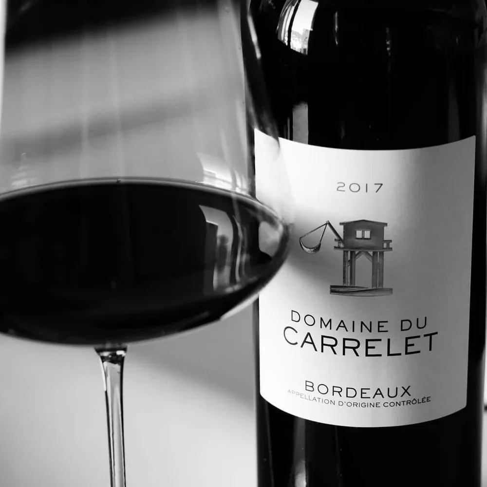 Bouteille du Domaine du Carrelet, millésime 2017 en AOC Bordeaux, accompagnée d’un verre de vin rouge, en noir et blanc.