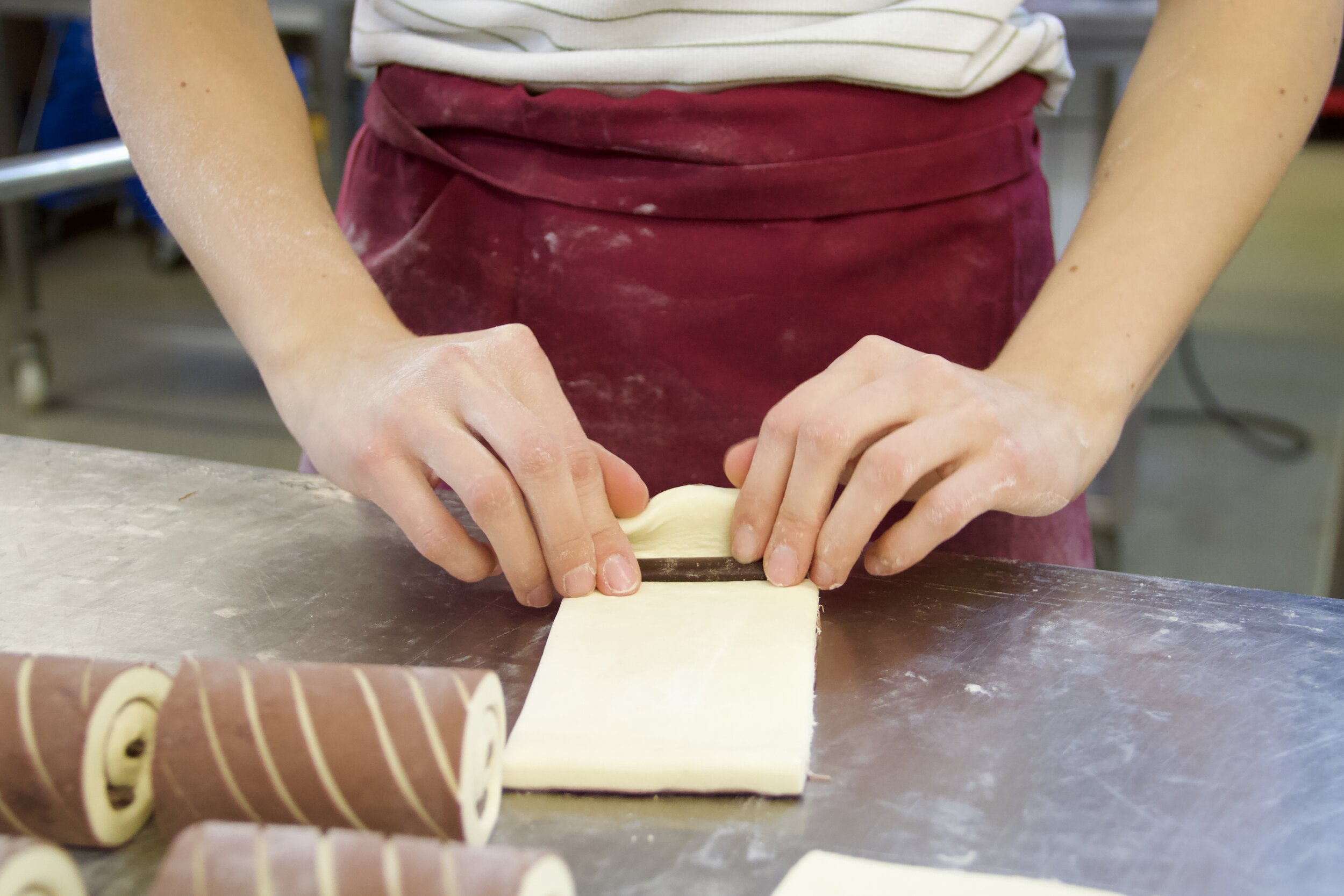 Eine Person rollt Teig mit einem Teigschneider auf einer Arbeitsfläche. Es liegen mehr eingerollte Teigrollen auf dem Tisch.
