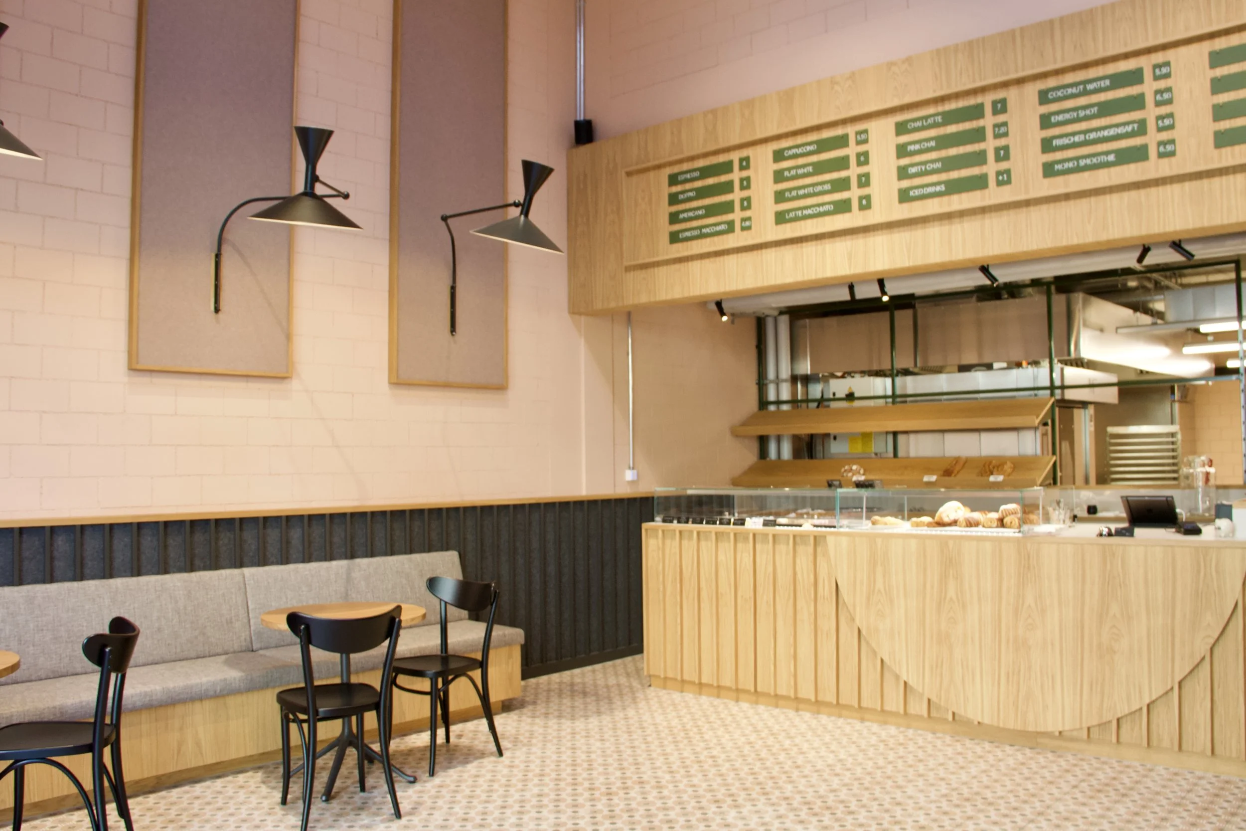 Innenansicht eines modernen Cafés mit Theke, Sitzbereich mit Stühlen und Tischen, helles Dekor mit Holzverkleidung und Wandlampen.