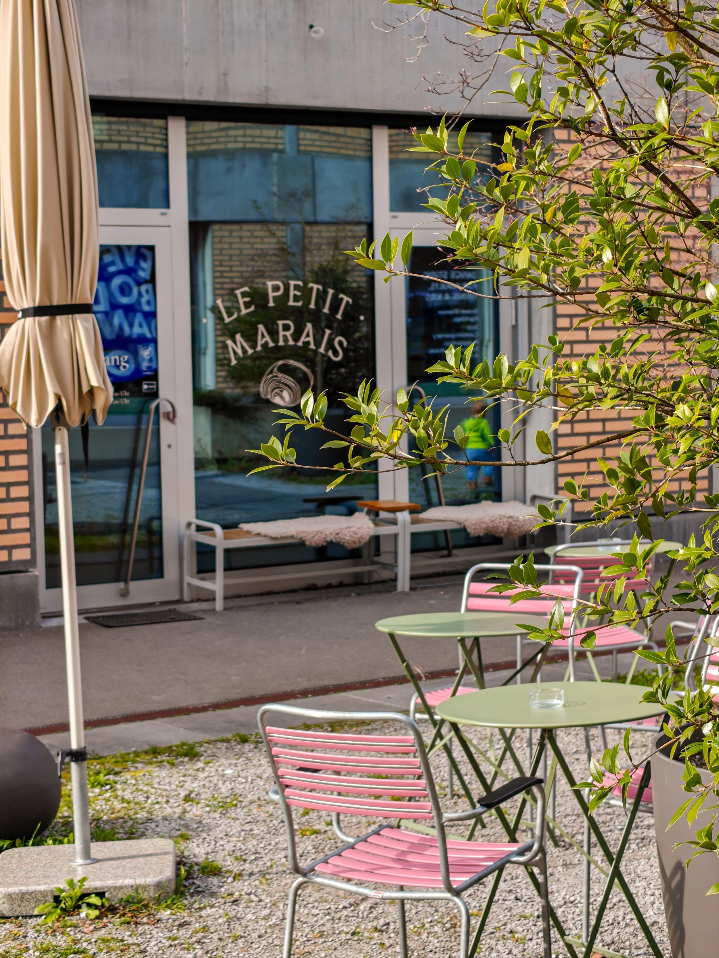 Ein Außenbereich vor einem Café mit grünen Tischen und pinkfarbenen Stühlen, umgeben von Pflanzen, mit einem Blick auf die Eingangstür zum Café "Le Petit Marais".
