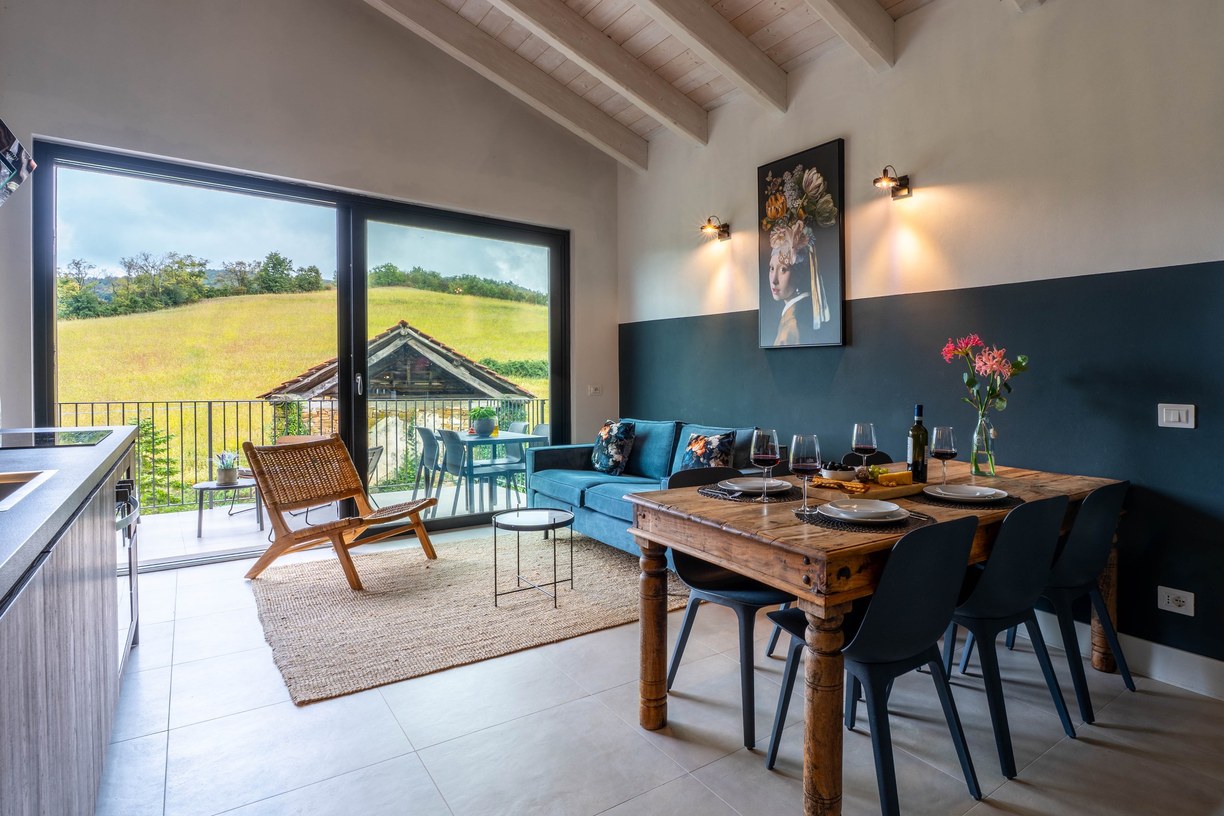 Gezellig interieur van een woonkamer met een grote glazen schuifdeur naar een balkon met uitzicht op groene heuvels, een blauwe bank, een houten eettafel met vier stoelen, en decoratieve elementen zoals een schilderij en bloemen.
