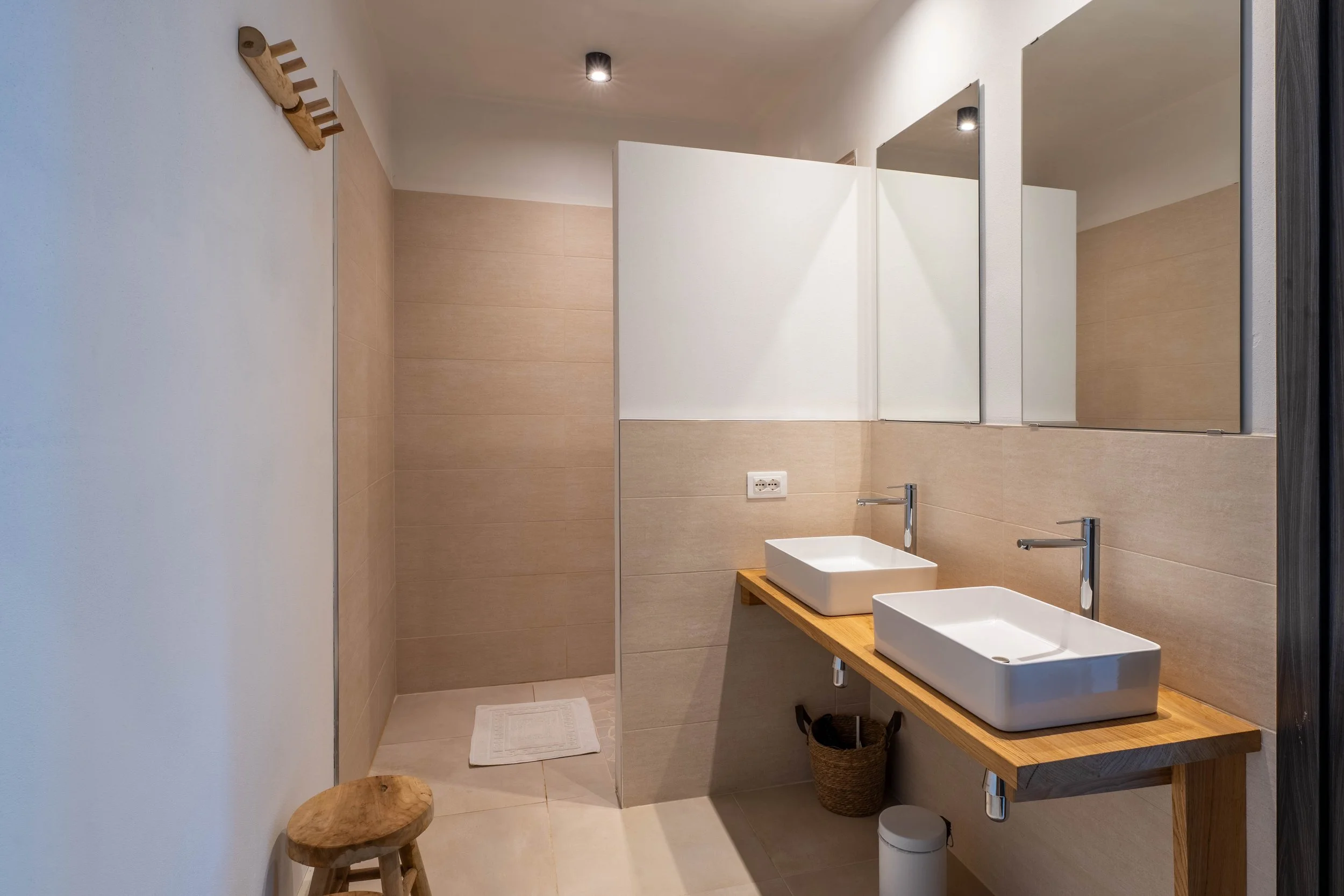 Moderne badkamer met dubbele wasbakken, houten blad, grote spiegels, beige wandtegels en een aparte inloopdouche met wit mattenloper.