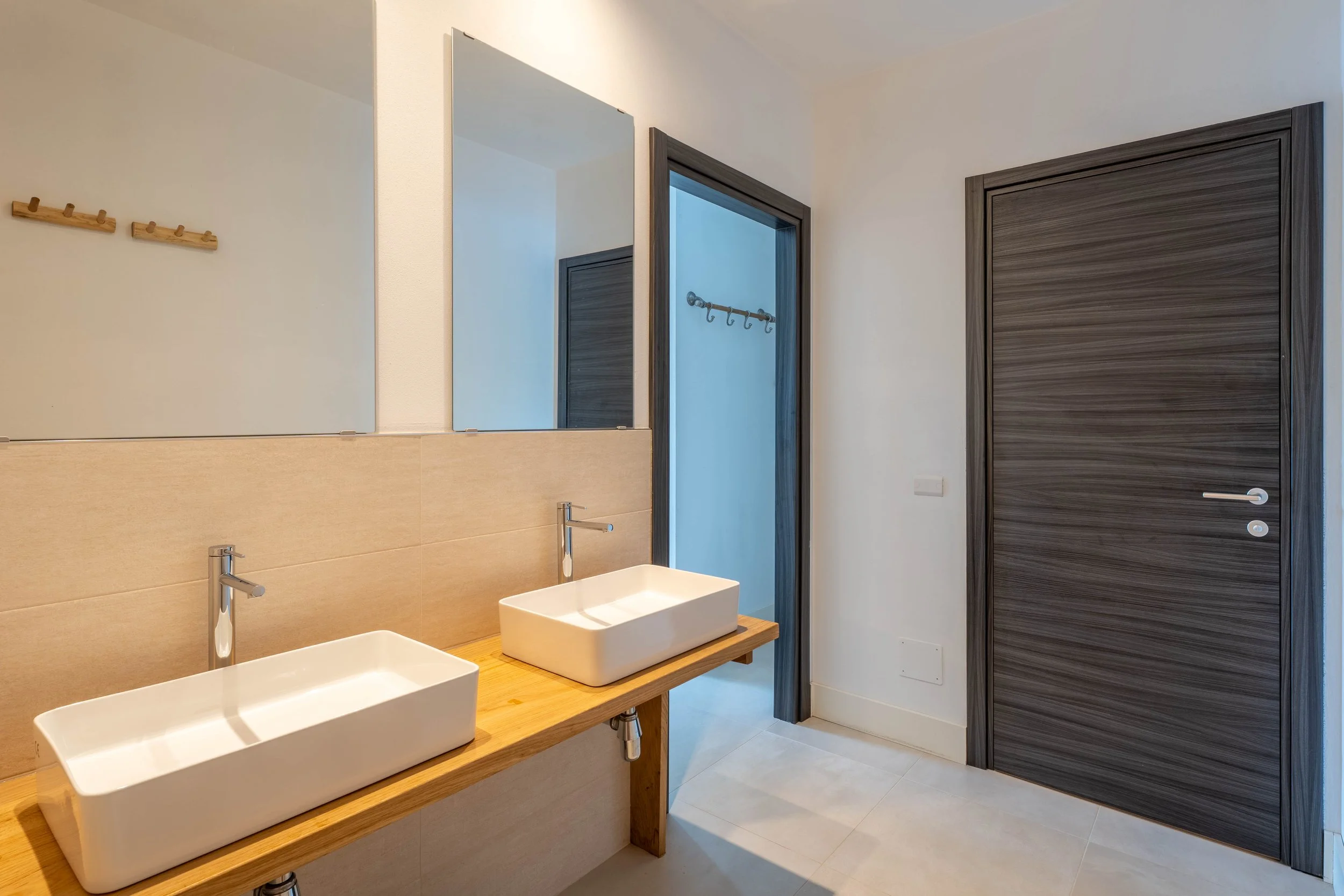 Badkamer met dubbele wastafel, grote spiegel, houten wastafelondersteuning, twee witte rechthoekige wasbakken, twee kranen, deur van donker hout, witte muren, lichte vloer.