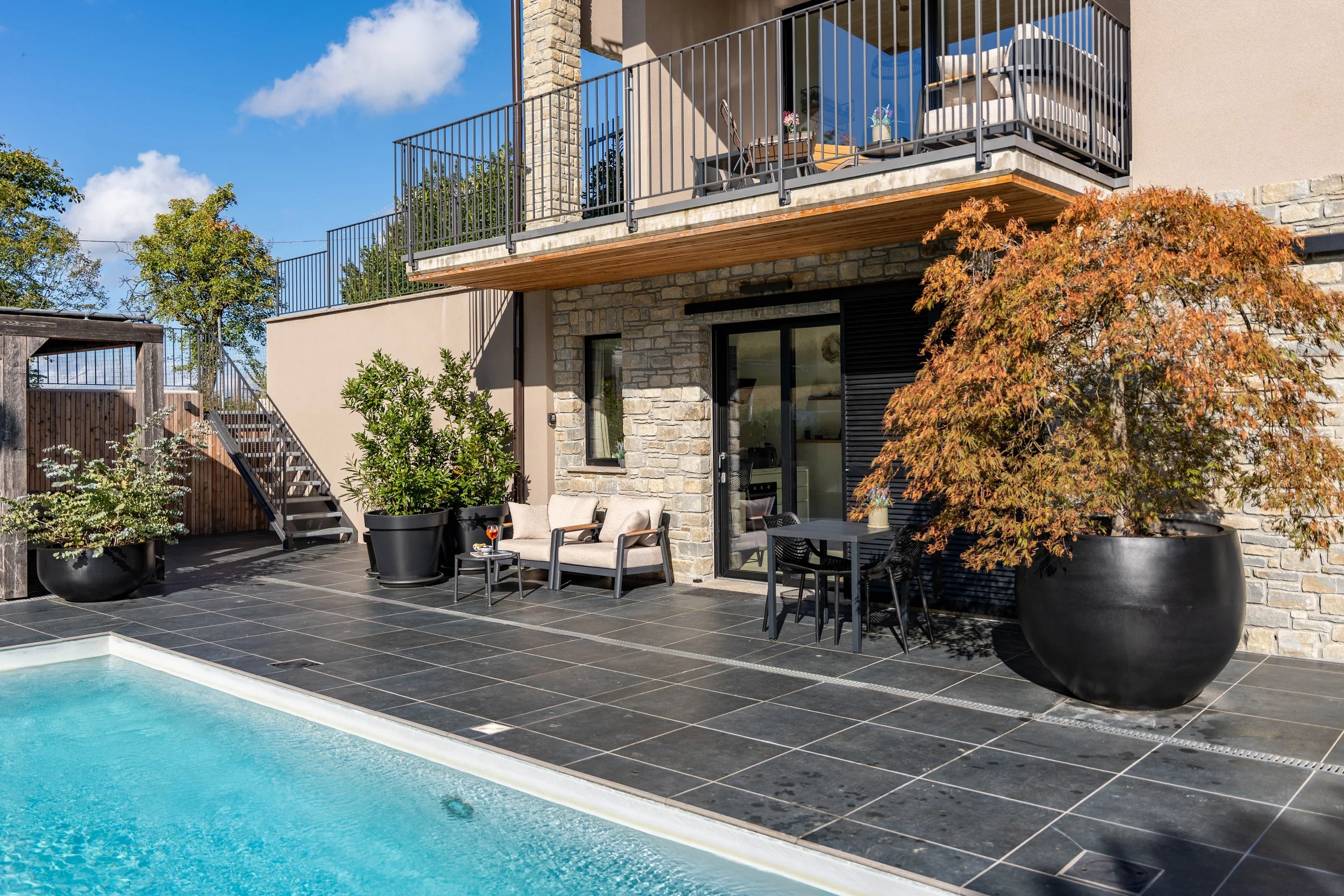 Terras van een modern huis met zwembad, grote plantenpotten, tuinmeubilair en een balkon.