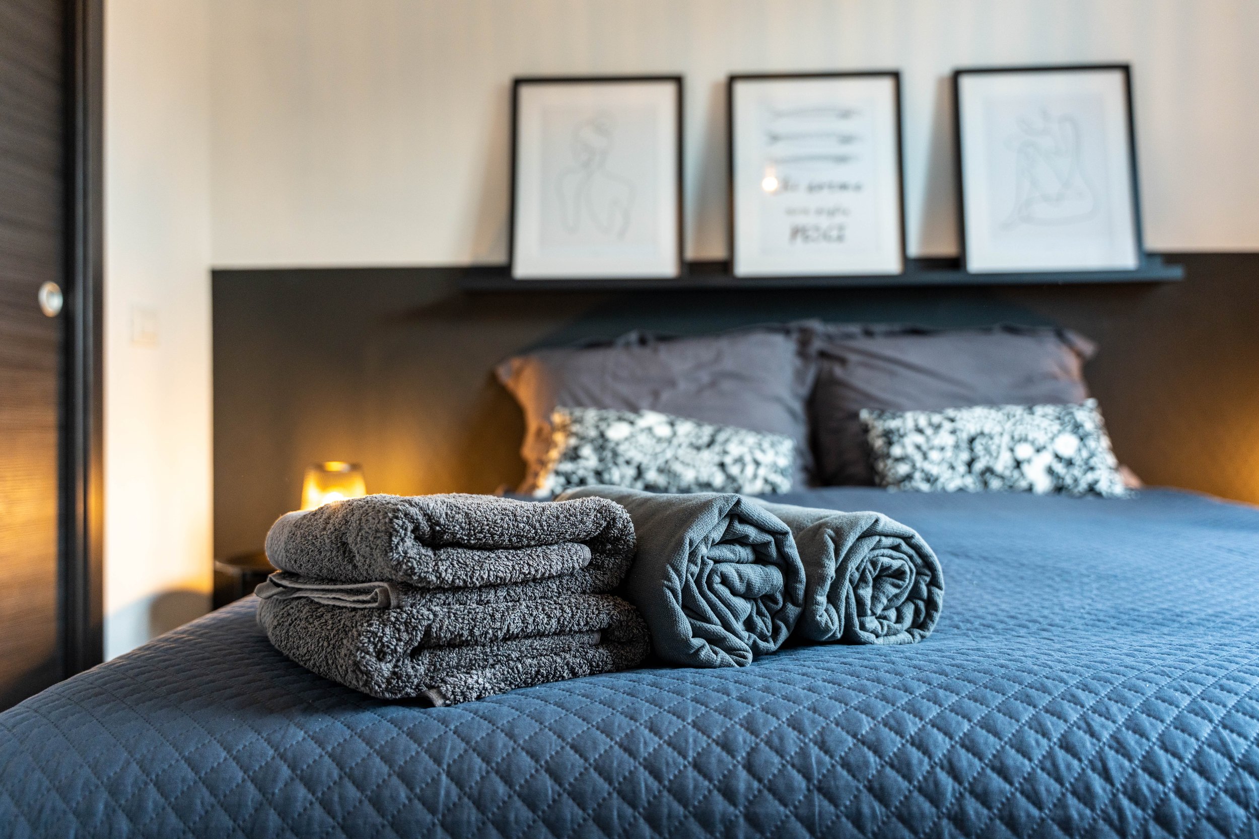 Hoofdeinde van een bed met ingelagen badjassen en handdoeken, op de achtergrond muur met artikelen en fotolijsten.