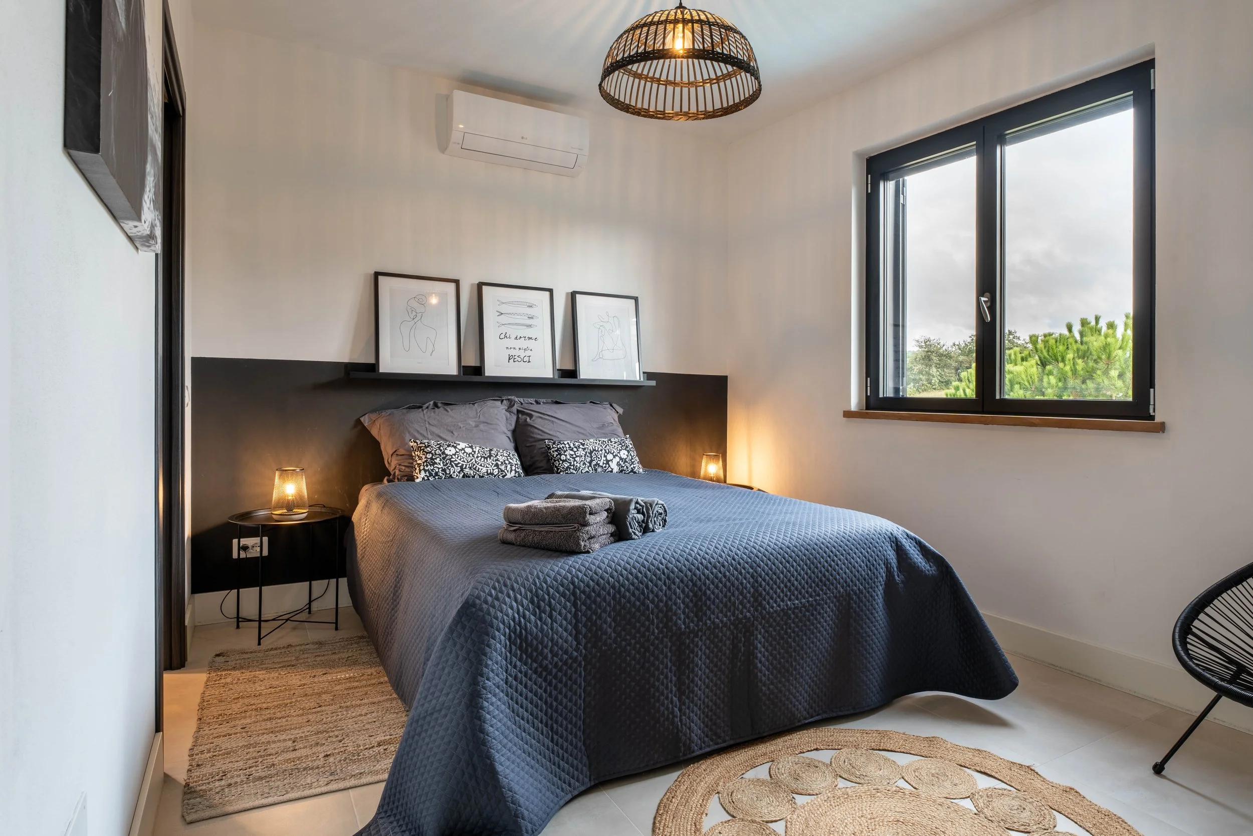 Een slaapkamer met een bed, versierd met zwarte en grijze kussens en een donkerblauw/dekbed. Boven het bed hangen drie zwart-wit kunstwerken in lijsten. Aan de linkerzijde bevindt zich een zwart nachtkastje met een klein lampje. Er liggen op het bed drie opgevouwen handdoeken of kussens. Er is een raam met een uitzicht op groen bomen en een lucht met wolken. Een licht gekleurd, rond vloerkleed ligt op de vloer, en er is een zwart, ronde stoel rechts in beeld.